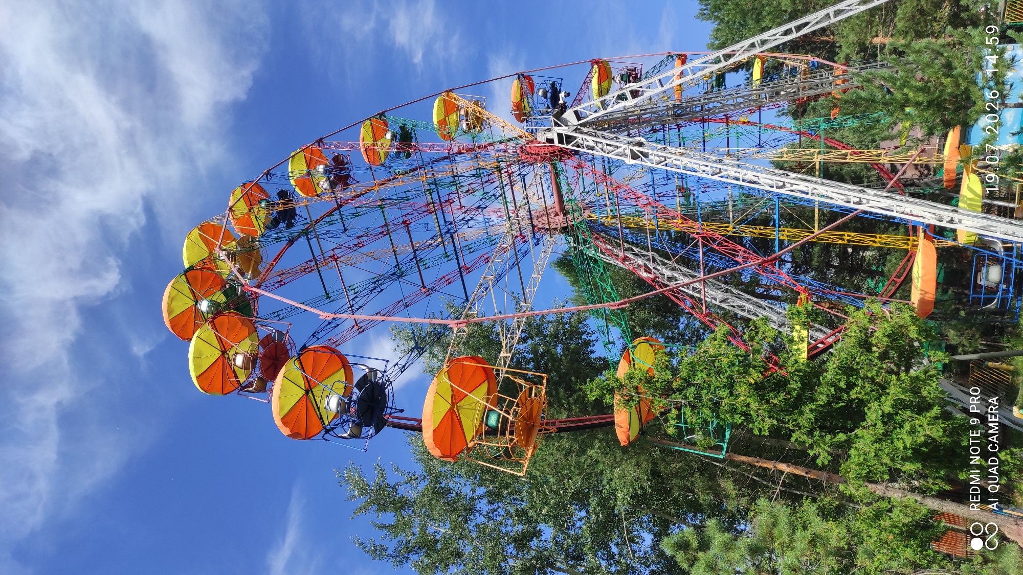This image captures a vibrant, colorful Ferris wheel against a bright blue sky with scattered white clouds. The Ferris wheel is a prominent amusement park ride, featuring numerous gondolas, each adorned with yellow and orange canopies. The structure itself is a complex lattice of metal beams painted in various bright colors, including red, blue, yellow, and green, adding to its playful and festive appearance. Trees with lush green foliage surround the base of the Ferris wheel, indicating an outdoor setting, possibly a park or an amusement area. The lower portion of the image shows some greenery and hints of other structures or ground, suggesting a public space. A watermark at the bottom left corner reads "REDMI NOTE 9 PRO AI QUAD CAMERA" and at the bottom right "19.07.2026 14:59". While the image does not explicitly state the location in Chaikovsky, Russia, it depicts a scene typical of a town's recreational facilities or a local park during fair weather.