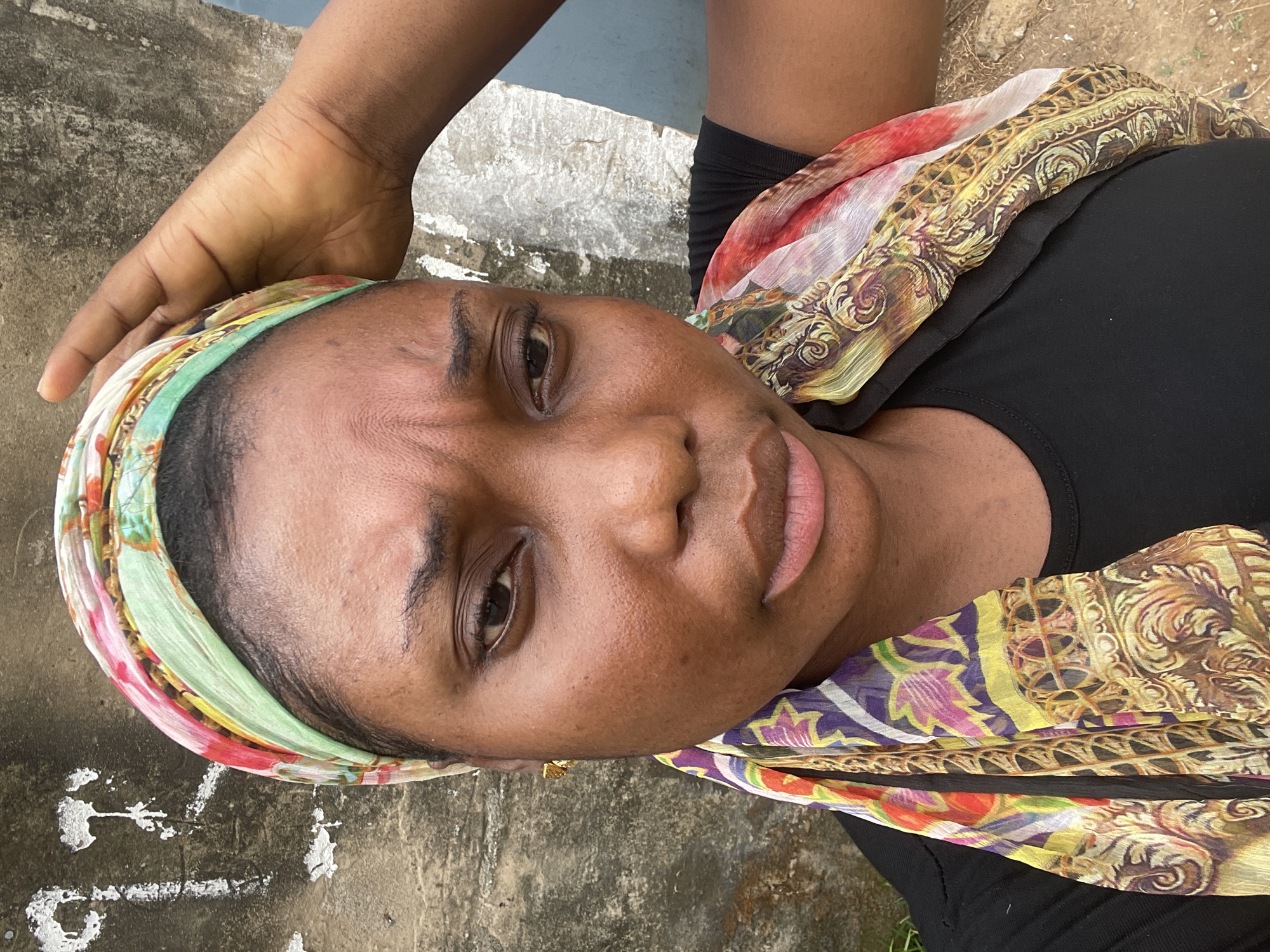 The image shows a close-up of a woman's face, positioned at an angle. She has dark skin and is wearing a colorful headscarf. A hand rests on her forehead. She is wearing a black top, and a patterned scarf is draped over her shoulder. The background appears to be a concrete surface with some markings. The location is in Abuja, Nigeria.