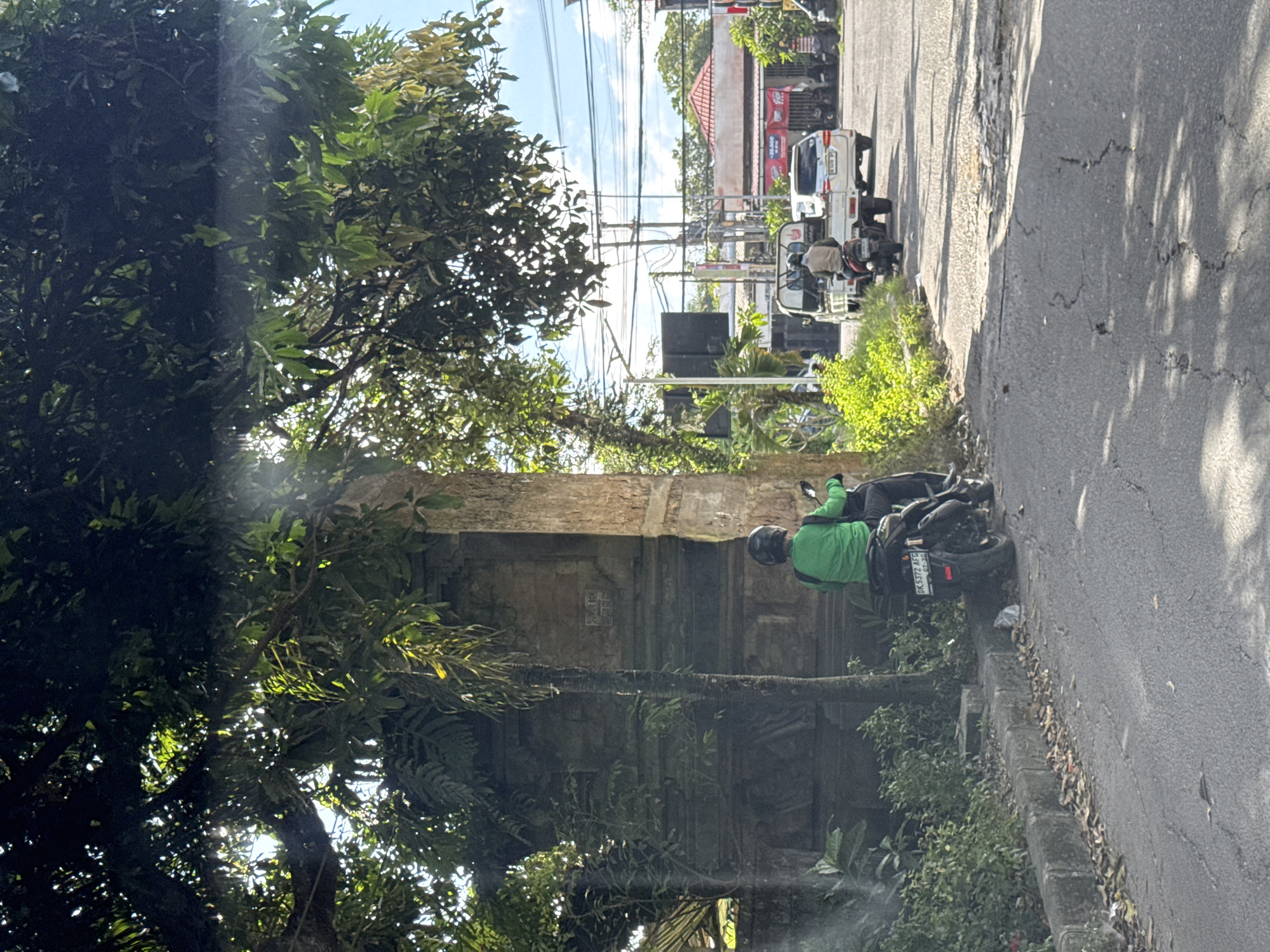 A horizontal outdoor shot taken from a slightly elevated position shows a street scene in Kuta Selatan, Indonesia. Along the right edge of the image, a cracked asphalt road stretches into the distance. A scooter parked on the shoulder has a rider wearing a bright green shirt and helmet. The license plate reads “DK5372 AFC.” To the left of the scooter, a mossy stone structure is partially obscured by a large tree. In the background, vehicles and buildings are visible under a blue sky. Overhead power lines crisscross the scene. Sunlight filters through the trees, creating bright spots on the road and foliage.
