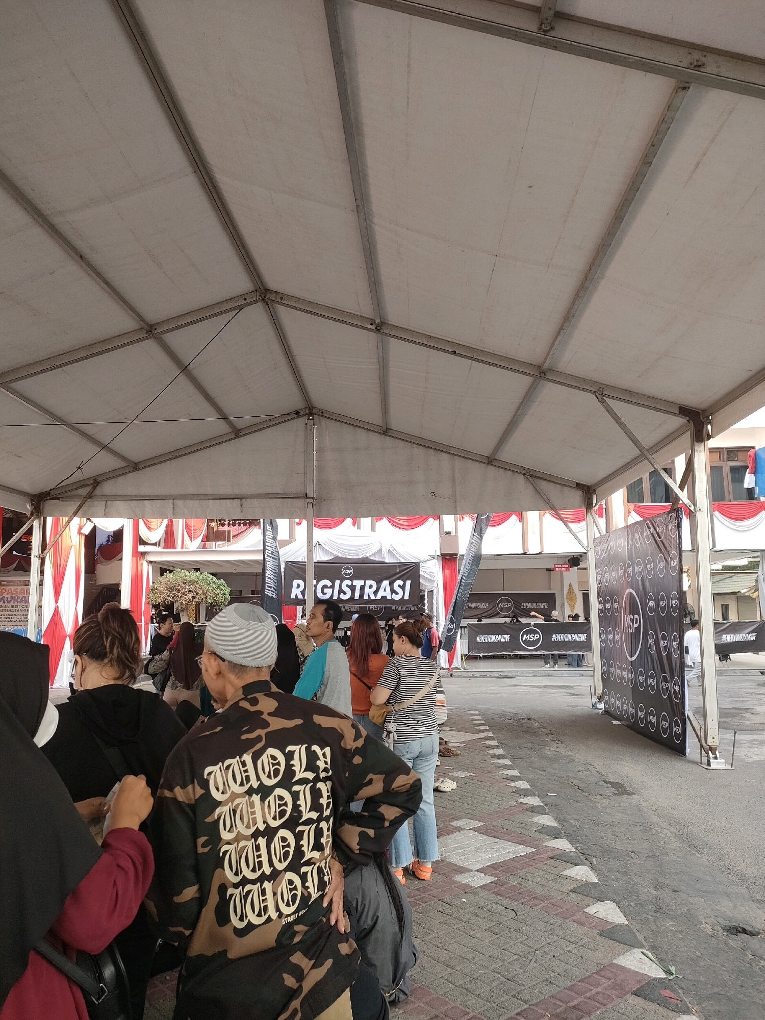 An outdoor registration area under a white tent is depicted. A line of people, seen from behind and the side, are waiting to register. The tent has metal supports and a white fabric roof. In the background, a banner with the word "REGISTRASI" is visible, along with red and white decorative drapes. Further back, a building facade can be seen with windows. To the right, a large black banner with circular logos is displayed. The ground is paved with a combination of bricks and asphalt. The image appears to have been taken on a bright, possibly overcast day. The location is identified as Banjarmasin, Indonesia.