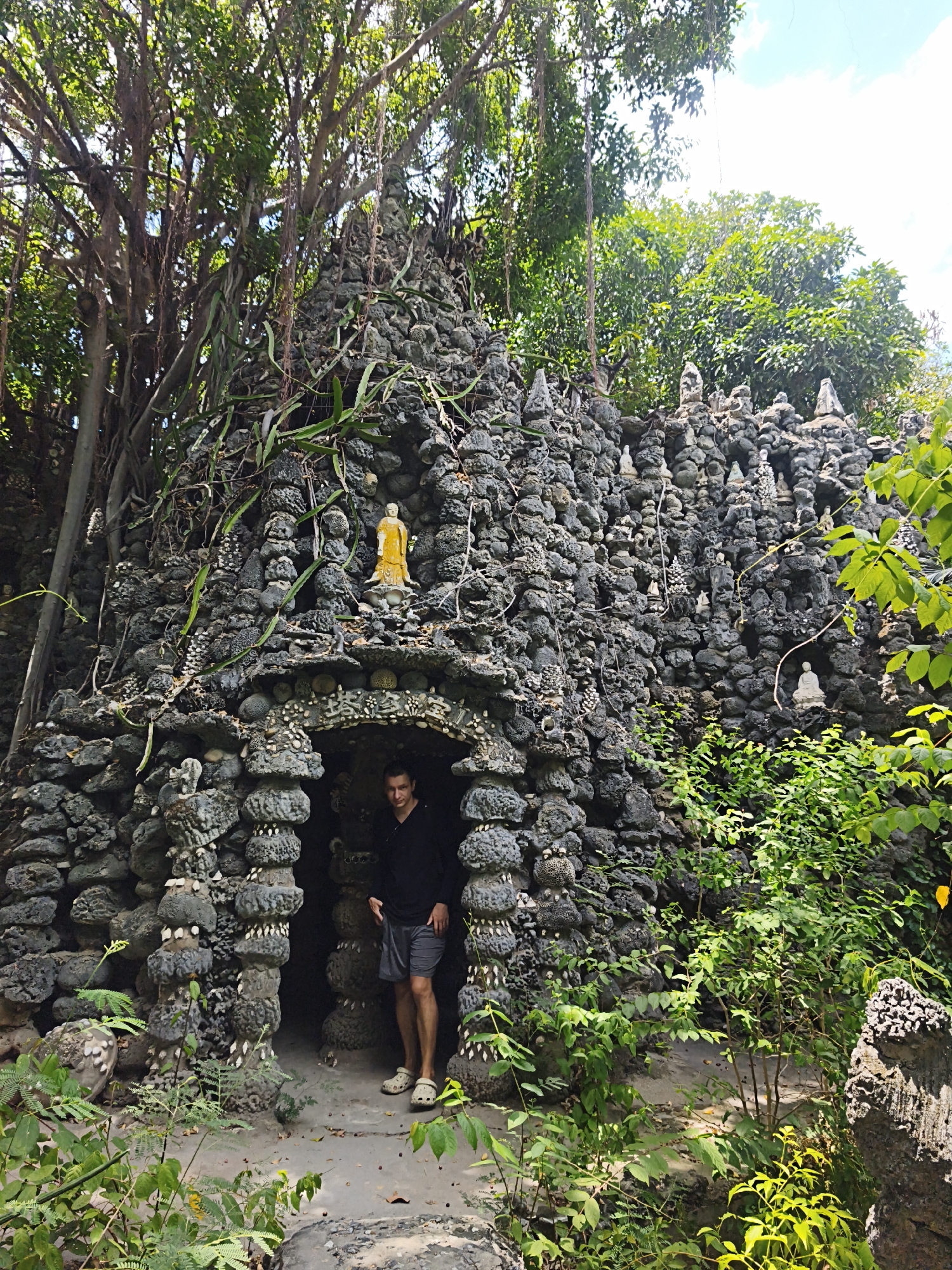 A man stands in the arched entrance of a rustic stone structure overgrown with vines and foliage. The structure, appearing to be a small temple or shrine, is constructed from dark, irregular stones and is adorned with numerous small statues of Buddhas and other figures. A larger golden Buddha statue is visible on top of the structure. The scene is set within a lush, natural environment, suggesting a tropical or sub-tropical location. The man is wearing a black long-sleeved shirt and grey shorts, with light-colored clogs on his feet. The overall impression is one of an ancient, somewhat hidden spiritual site. This site is likely the "Thien An Ton Pagoda" in Cam Ranh City, Khanh Hoa Province, Vietnam, known for its unique stone sculptures and overgrown landscape.