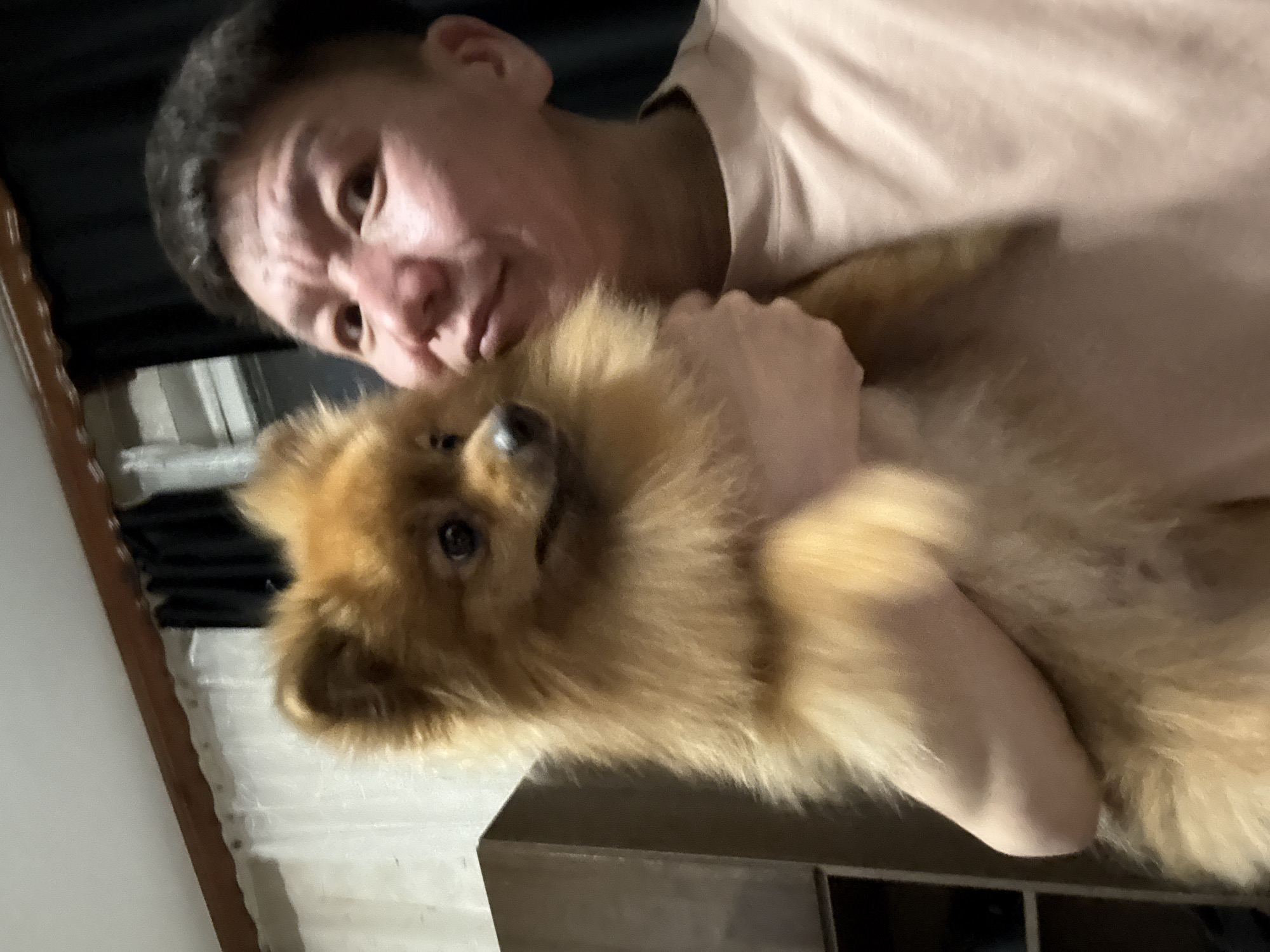A person, with short dark hair and light skin, is holding a small, fluffy, light brown dog. The person is looking towards the camera and smiling slightly. The dog is facing left and looking away from the camera. They are indoors, and the background includes a wooden cabinet on the left and curtains on the right. The lighting appears to be artificial. The image was taken in Khimki, Russia.