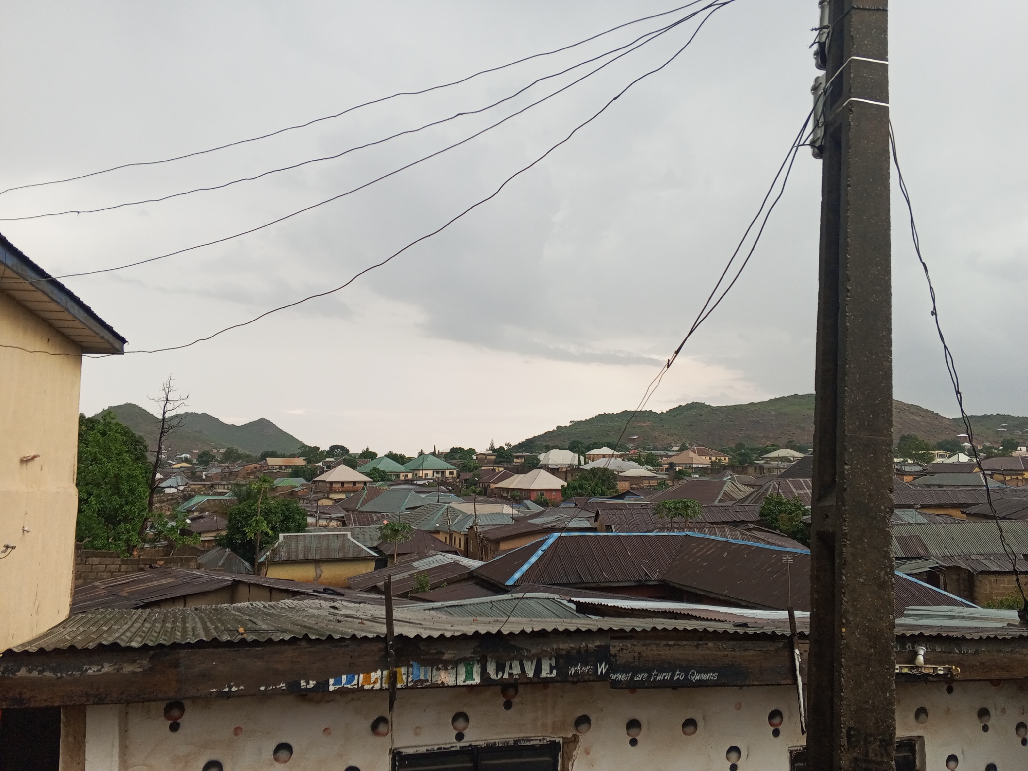 The image depicts an overcast urban scene in Gwong, Nigeria. In the foreground is a textured, weathered concrete wall with round cutouts and faded text that appears to read, "...CAVE where Women are turn to Queens." Above the wall, a dense cluster of rooftops varies in color and material, primarily corrugated metal, with a few trees interspersed. The background reveals low, green-covered hills under a grey sky. An aging concrete utility pole dominates the right side of the frame, with multiple electrical wires stretching across the sky. The setting suggests a lower-income residential neighborhood. No people are visible.