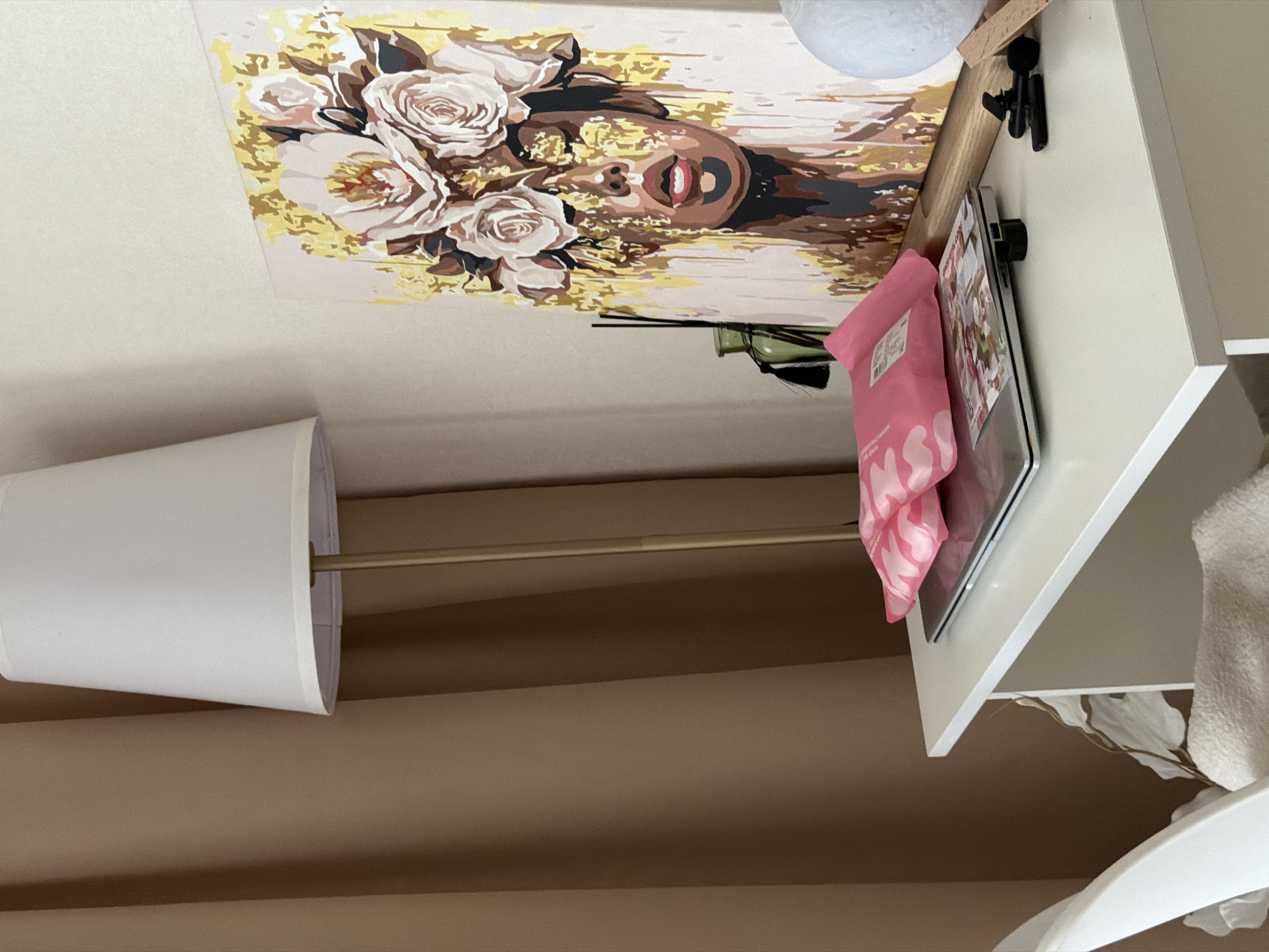 The image shows a white desk against a wall. On the desk, there is a laptop, two pink packets, a framed painting of a woman with flowers on her head, and a green bottle with reeds. To the left of the desk, there is a tan curtain and a floor lamp with a white shade. A white chair is partially visible to the left of the desk. The scene is set indoors, likely in a bedroom or living area, but there is no specific indication of the city of Semiluki, Russia.