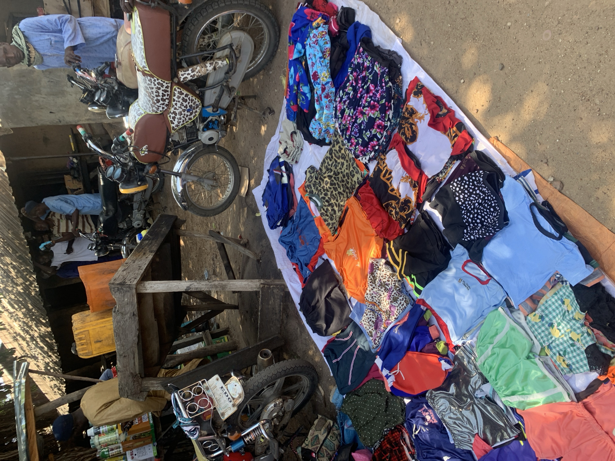 The image depicts an outdoor market scene in Nigeria, likely in the morning or late afternoon given the dappled sunlight and shadows. The foreground features a spread of colorful clothing laid out on a white cloth, interspersed with more items on the ground. The clothing includes a variety of patterns and colors, such as floral prints, leopard print, polka dots, and solid bright hues like orange and green.
Behind the clothing display, several motorcycles are parked. One motorcycle, a brown and tan model with a distinctive patterned seat, is prominently featured. The motorcycles suggest a mode of transportation and potentially part of the market's setup.
In the background, there are structures that appear to be market stalls or simple buildings, constructed with corrugated metal roofing and wooden beams. Several individuals are visible, some appearing to be vendors or customers. One person in the upper left is wearing a light blue robe and head covering. Another person is leaning over a motorcycle in the middle left. Further back, other figures are less distinct.
On the left side of the image, several large yellow and orange plastic containers are stacked, possibly used for storage or as part of the market's goods. Some bottles and other items are also visible on a table-like structure. The overall scene conveys a bustling, informal market environment. No specific text is clearly legible in the image.