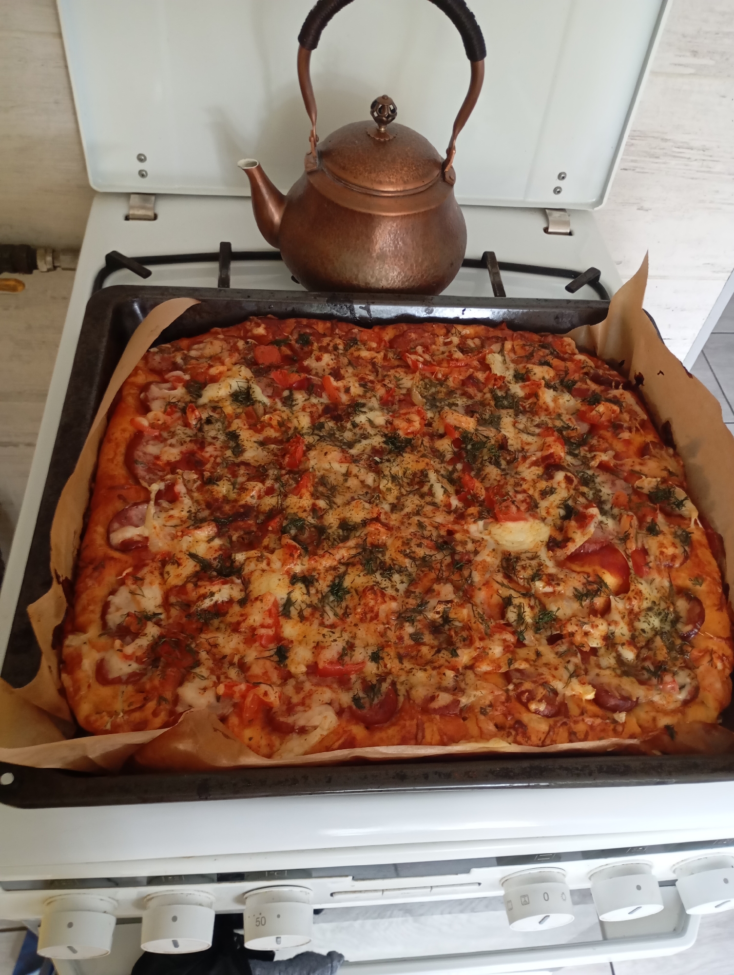 The image shows a rectangular pizza on a black baking sheet lined with parchment paper, sitting on a white stove. The pizza is topped with tomato sauce, cheese, sausage slices, diced tomatoes, and herbs, possibly dill. Above the pizza is a copper-colored kettle with a dark handle, placed on the stove's burners. The stove is located in a kitchen with white cabinets. The specific location within the city of Bydgoszcz, Poland cannot be determined without further information or context from the image.