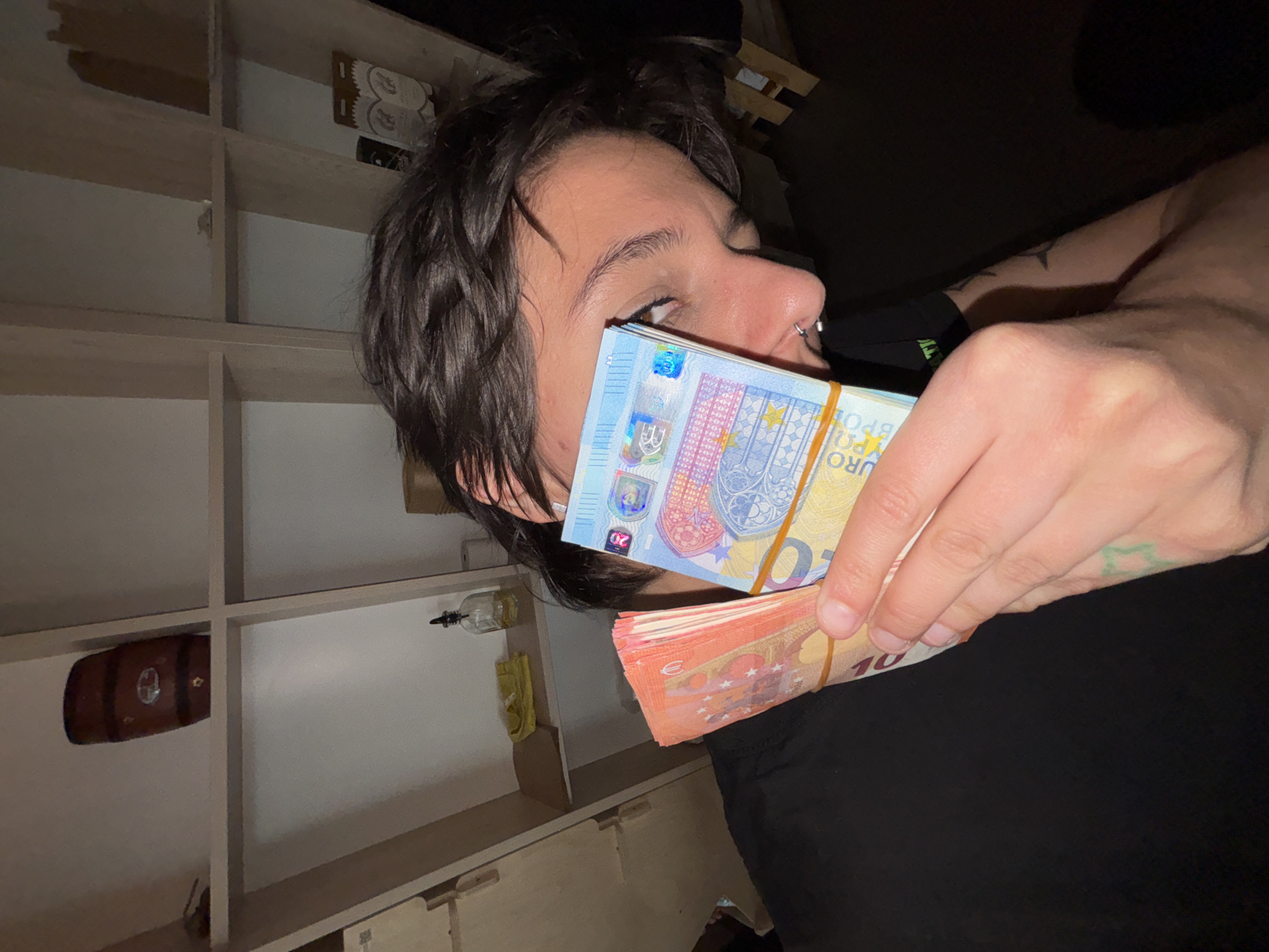 An indoor scene, likely set in Sofia, Bulgaria, features a person holding two stacks of Euro banknotes under dim lighting, suggesting evening or night. The subject is seen from the right side, revealing dark hair, an eye, nose, and forehead. A silver septum piercing is visible on their nose. Their left arm and hand hold the bundled money close to their face.
One stack of banknotes is primarily blue and green, consisting of 20 Euro notes. "EURO" and the denomination "20" are clearly visible, along with security features and text like "BCE ECB EZB EKT EKP". This stack is secured with a light brown rubber band. Below it, a second stack of red and orange 10 Euro notes is held, also bound by a rubber band, with "EURO" and "10" visible.
The background shows a light-colored shelving unit or bookcase with multiple compartments. Most shelves are empty, but some items are present: a small, dark wooden barrel on one shelf, a clear glass bottle with a black pourer on another, and some cardboard packaging on a top shelf. The overall setting appears casual, possibly a home or a storage area. The focus of the activity is the display of the two bundles of cash.