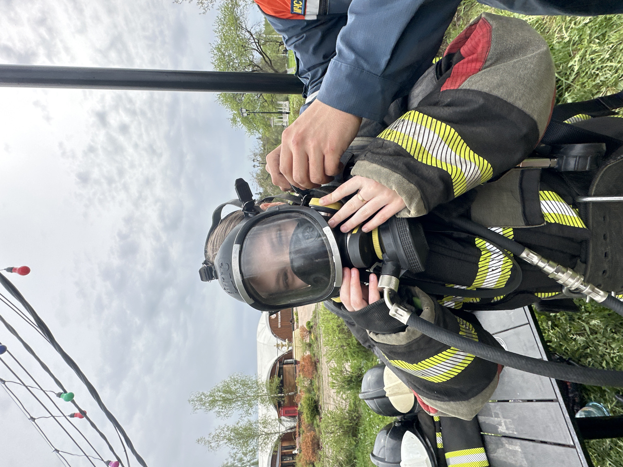 A person wearing a firefighter’s protective suit and mask is being assisted with the mask’s adjustment by another person wearing a blue uniform with an orange and white patch. The location is Grebnevo, Russia. A building with a distinctive arched roof is visible in the background, along with trees and grass. String lights with colored bulbs are partially visible on the left side of the image. A black pole is visible in the upper left corner. The individuals are situated on a surface that appears to be wooden planks.