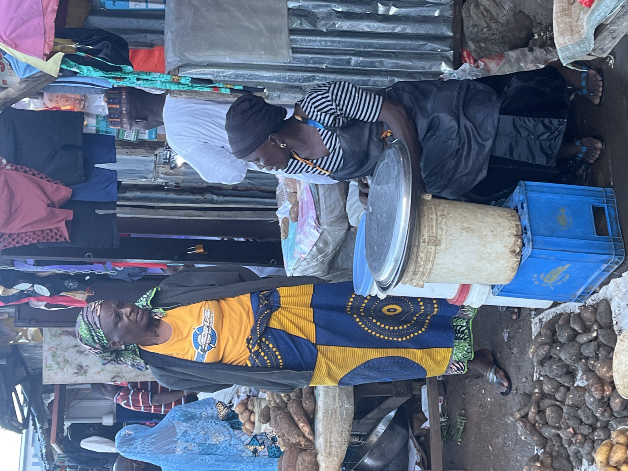 In the city of Gura Top, Nigeria, a bustling marketplace scene unfolds. Two women, presumably vendors, engage in commerce. The woman on the left, draped in a vibrant yellow and blue patterned skirt and a green headscarf, stands next to a display of root vegetables. She wears a dark jacket over an orange t-shirt. To her right, another woman, wearing a black and white striped shirt, a black headscarf, and a black apron, sits behind a large metal platter and bucket. She appears to be attending to customers or her stall. In the background, other market-goers, stalls with hanging clothing, and more produce are visible, suggesting a lively and active trade environment. The overall impression is one of daily life and economic activity in a Nigerian marketplace.