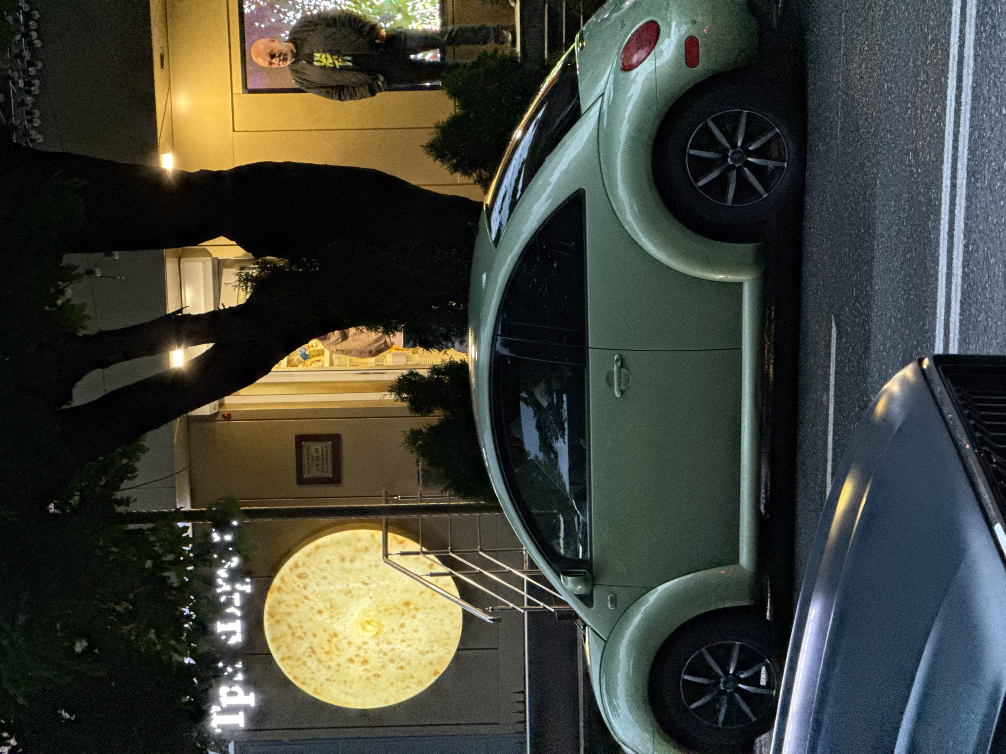 A light green Volkswagen Beetle is parked on a street at night, alongside a dark gray car. The Beetle is oriented vertically in the frame, with its wheels visible. To the left of the Beetle is a building with a lit-up circular sign and a framed plaque on the wall. A person is partially visible in a window of the building above the Beetle. The scene appears to be outdoors, possibly in a town or city, given the presence of streetlights and pavement. The overall atmosphere is dark, typical of nighttime. There is some foliage on the left side of the image, suggesting the presence of trees or bushes. The text "tp. t. di." is visible vertically in white lettering on the left side of the image, partially obscured by foliage and possibly a string of lights. The framed plaque on the wall has text on it, but it is too small to be read.
