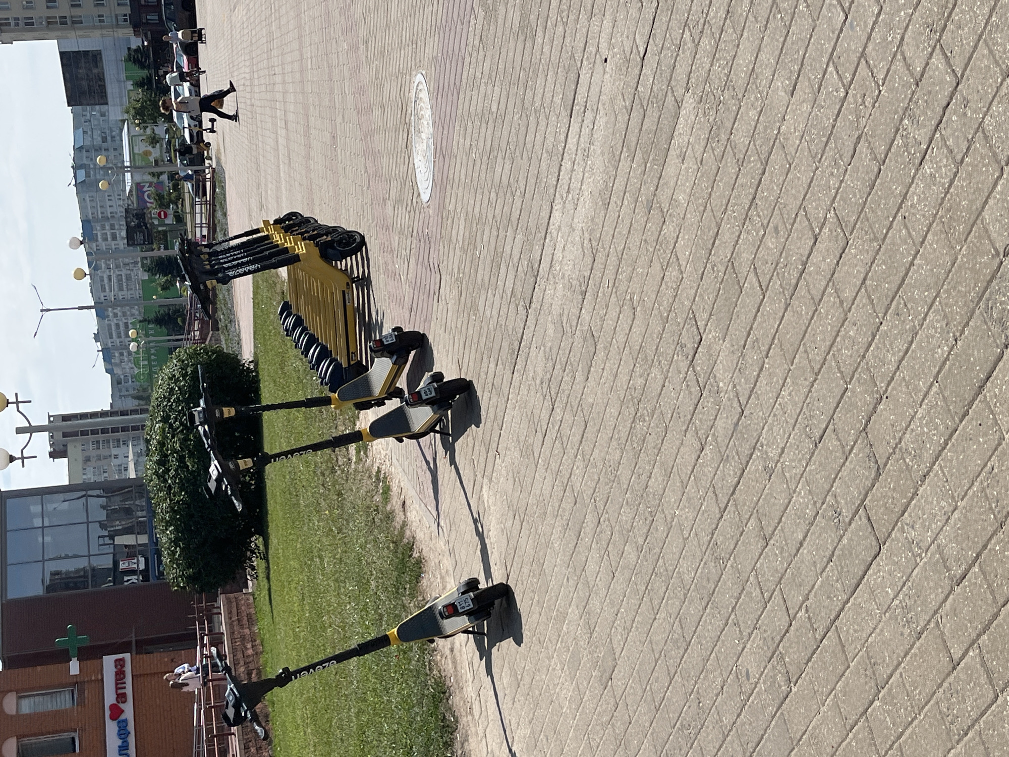 The image depicts an outdoor scene in Minsk, Belarus, featuring a row of parked electric scooters lined up along a paved walkway. In the background, a building with a brick facade and glass windows is visible, along with a green cross sign and lettering that appears to be Cyrillic. Further in the distance, apartment buildings and trees suggest an urban environment. Several individuals are present, with one person walking across the paved area and another in the background near a vehicle. The scooter charging station is yellow and black, and individual scooters have license plates.