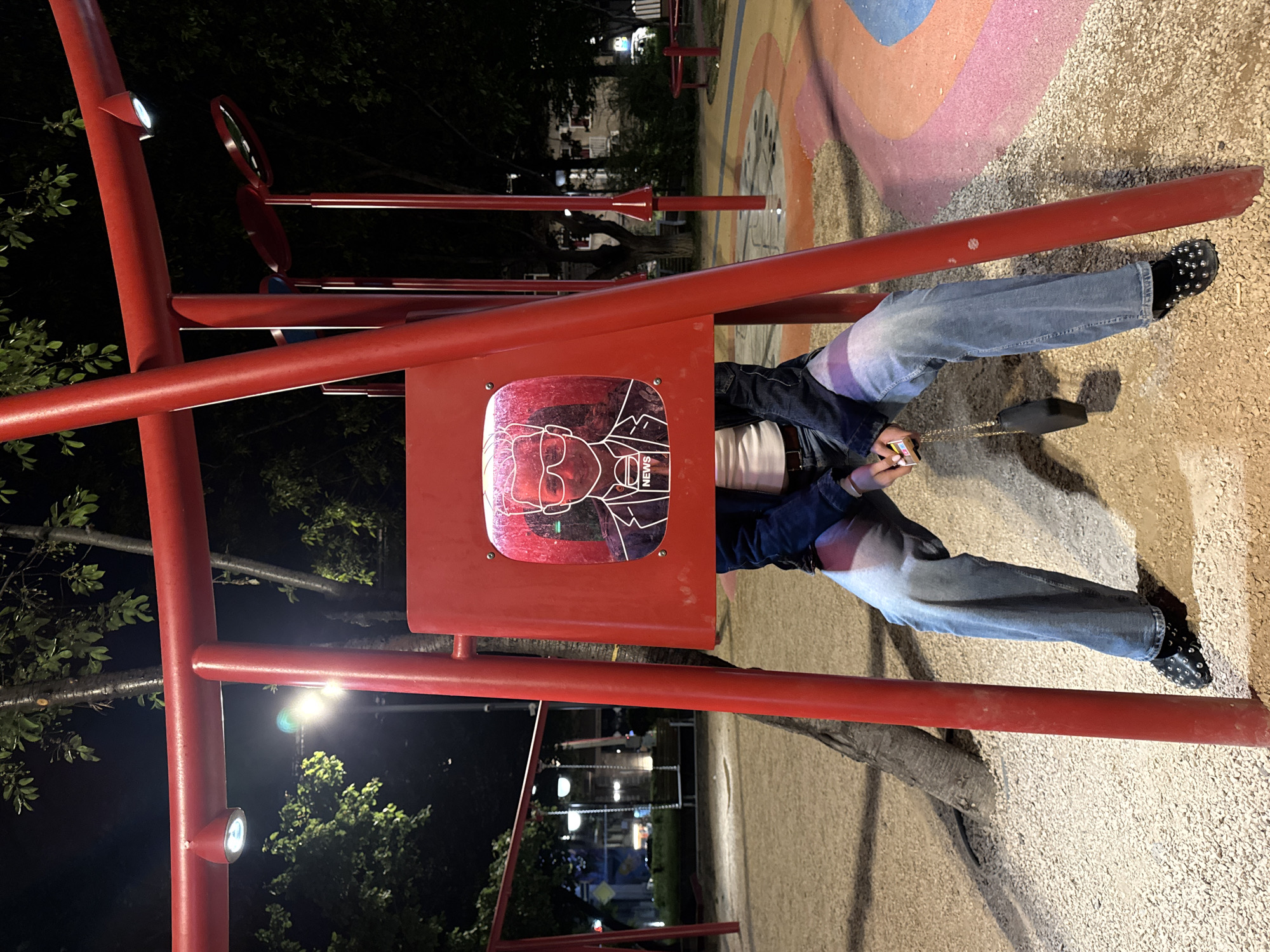 The photograph was taken at night in Yerevan, Armenia. It features a red metal playground structure with a small sign containing a graphic of a person's face and the word "NEWS." The structure is situated in a gravel and painted surface area. A person, wearing jeans and black shoes with studs, is using the structure. The background is blurred, but trees and city lights are visible.