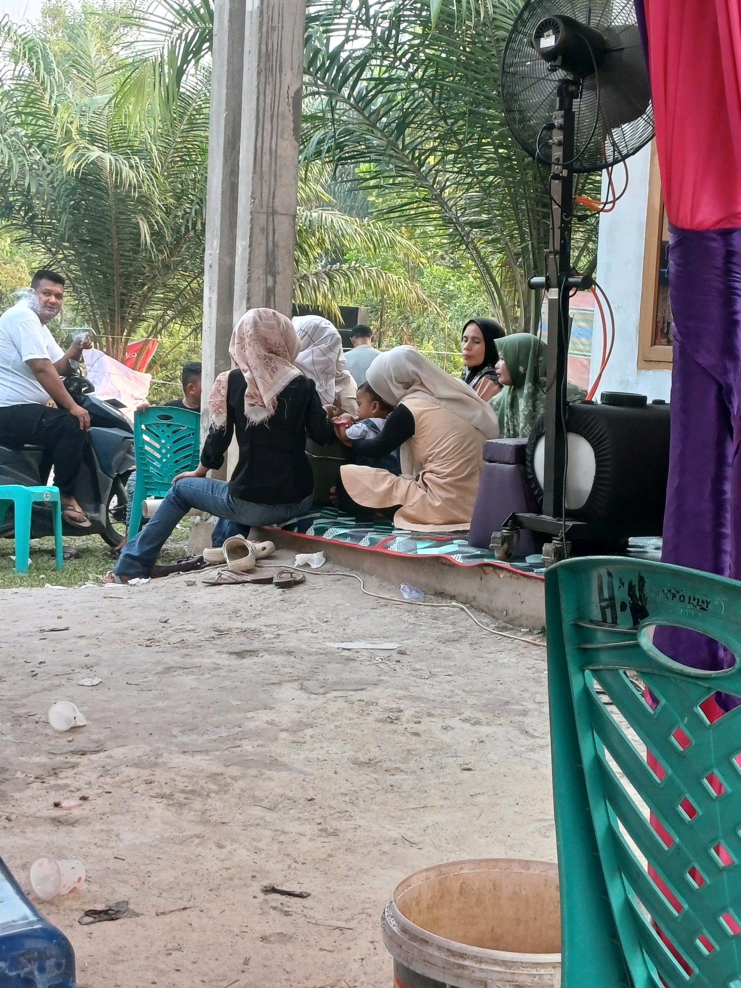 This image depicts an outdoor gathering in Dumai City, Indonesia, likely a social event such as a wedding reception or community meeting, given the presence of decorations and a communal seating arrangement. The setting appears to be a casual outdoor space, possibly a yard or a public gathering area, with palm trees visible in the background, indicative of a tropical climate.
In the foreground, several individuals are seated on the ground or on chairs. A man in a white t-shirt is seated on a scooter on the left, engaged in conversation and holding a cigarette. To his right, a group of women and a child are gathered. One woman, wearing a black top and jeans, is seated with her back to the camera, looking towards the child. Another woman in a light-colored headscarf is holding a baby, interacting with it. A third woman in a green headscarf is also visible, facing the group. The women are dressed in traditional Islamic attire, with headscarves and modest clothing.
In the middle ground, a large industrial fan is positioned, suggesting a need for cooling in the warm climate. Colorful draped fabric, in shades of pink and purple, is visible on the right side of the image, likely part of the event's decorations. A black speaker or equipment box is also present near the fan. Various chairs, including a green plastic chair in the foreground, are scattered around the scene.
The ground is a mix of dirt and concrete, with some scattered debris such as plastic cups and a discarded flip-flop. The overall impression is that of a lively but relaxed community event taking place in a semi-urban or rural setting in Dumai City. The presence of the fan and the attire of the people suggest the weather is warm and humid. The scene captures a moment of social interaction and shared activity among the residents.