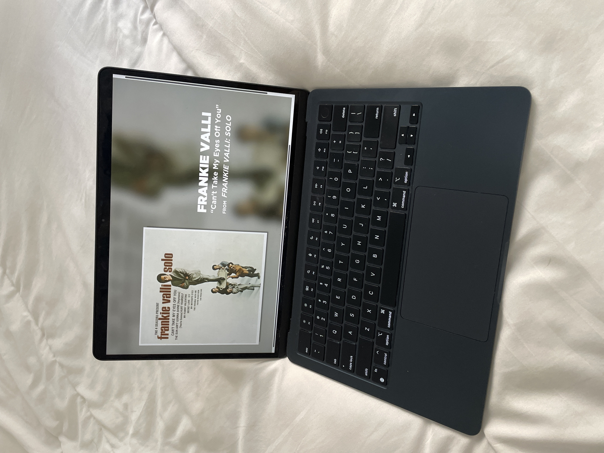 The image shows a laptop open on a white bedsheet. The screen displays an album cover of Frankie Valli: Solo, presenting the songs "Can't Take My Eyes Off You", "The Sun Ain't Gonna Shine (Anymore)", "(You're Gonna) Hurt Yourself", "My Funny Valentine", "Secret Love IVY", "My Mother's Eyes", "The Trouble With Me", and "You're Ready Now". The keyboard is visible, along with the trackpad. The scene occurs in an interior environment with white bed sheets as a backdrop.