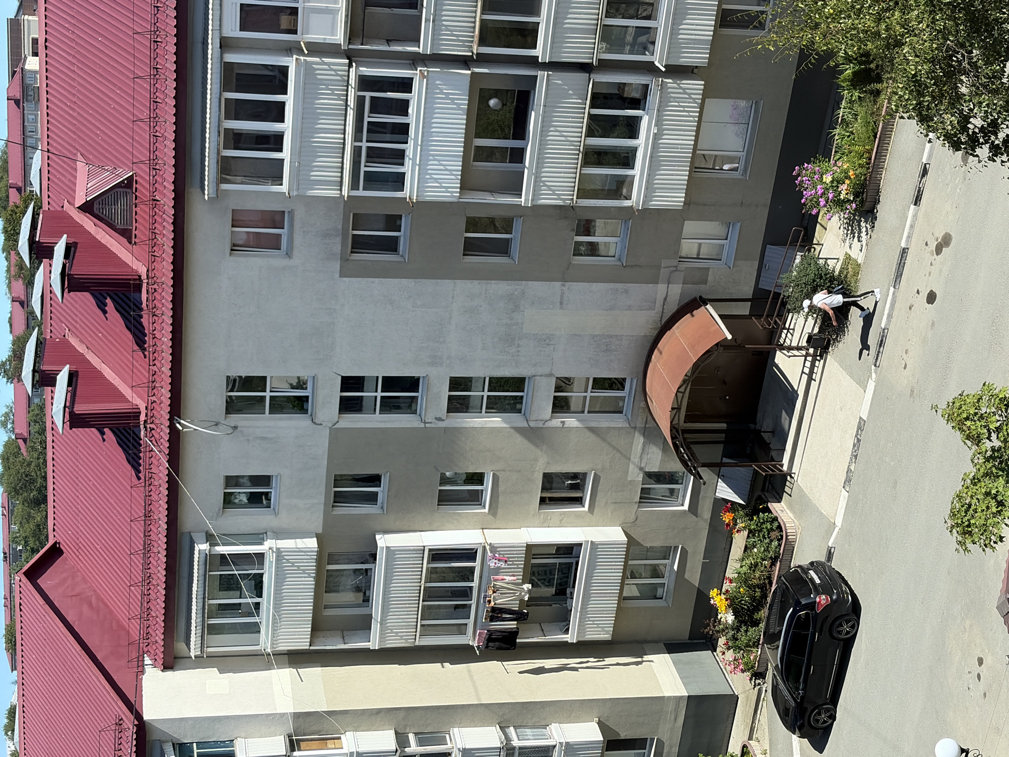 The image depicts a multi-story apartment building with a red corrugated metal roof, located in Korsakov, Russia. The building has a light-colored facade with numerous windows and balconies, some of which have white vertical railings and enclosed glass fronts. Laundry is hanging on a clothesline on one of the balconies. In front of the building, a black SUV is parked on the street. Adjacent to the building, there is a flower bed with colorful flowers. A person wearing a white cap, white tank top, and dark pants is walking on the sidewalk. A covered entryway with a dark brown awning leads into the building. Trees and other buildings are visible in the background. The scene appears to be set on a sunny day.