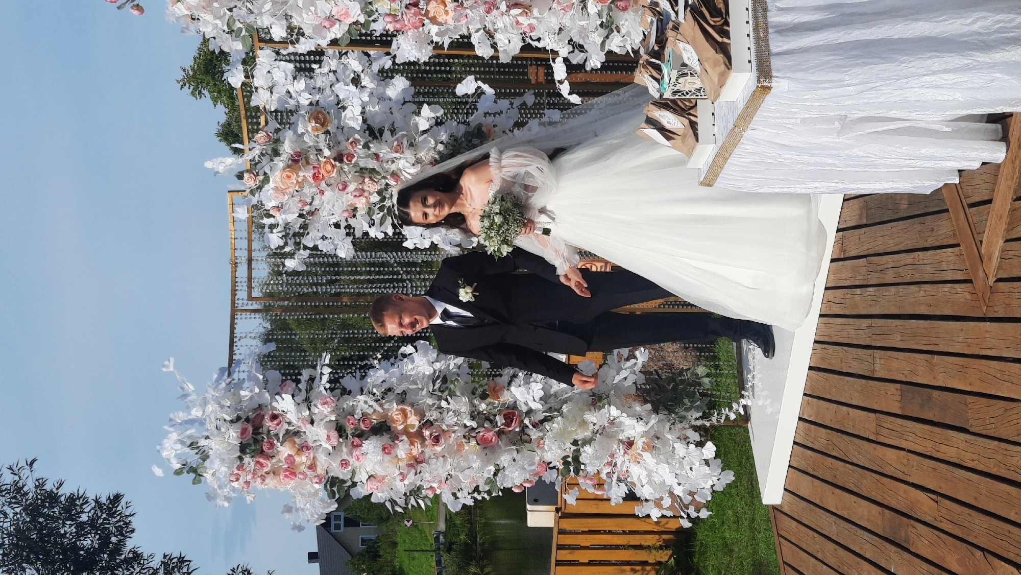 A wedding ceremony is taking place outdoors at the Жодзішкаўскі сельскі Савет, Belarus. A young couple, the bride and groom, are standing in front of a decorative wedding arch adorned with white and pink flowers and crystal strands. The groom, dressed in a black suit, white shirt, and black tie, stands on the left, smiling and looking to his left. The bride, in a white off-the-shoulder wedding dress with voluminous sleeves and a veil, stands on the right, holding a bouquet of white flowers and looking towards the camera. They are standing on a white platform, which is situated on a wooden deck. To the right of the platform, a table is set up with a white tablecloth, decorated with a gold beaded trim. On the table are several brown paper bags and decorative boxes. In the background, there is a body of water, a wooden fence, and lush green trees. The sky is a clear blue.
