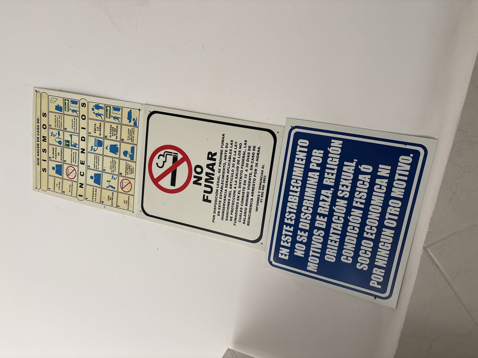 Three informational signs are displayed on a clean white wall in what appears to be an indoor public or commercial setting in Mexico City, Mexico. The image is taken at a slight angle, showing the signs neatly affixed to the wall.
The leftmost sign is a white rectangular panel with multiple numbered pictograms and Spanish text detailing emergency procedures. Titled "QUE HACER EN CASO DE: SISMOS" (What to do in case of: Earthquakes) and "INCENDIOS" (Fires), it provides instructions such as "CONSERVE LA CALMA" (Keep calm), "NO USE ELEVADORES" (Do not use elevators), "UBIQUESE EN ZONAS DE SEGURIDAD" (Locate yourself in safe zones), and "SI EL HUMO ES DENSO ARRASTRESE POR EL SUELO" (If smoke is dense, crawl on the floor).
The middle sign, also white, features a prominent red circle with a diagonal line through a lit cigarette, indicating "NO FUMAR" (No Smoking). Below, white text on a black background explains that smoking is legally prohibited and outlines penalties for non-compliance, citing "ARTICULO 30 DE LA LEY DE PROTECCION A LA SALUD DE LOS NO FUMADORES EN EL DISTRITO FEDERAL." It specifies fines based on minimum wage and potential arrest for repeat offenses, and provides a Mexican complaint hotline: "01 800 966 3863".
The rightmost sign is blue with bold white capital letters. It states: "EN ESTE ESTABLECIMIENTO NO SE DISCRIMINA POR MOTIVOS DE RAZA, RELIGIÓN ORIENTACIÓN SEXUAL, CONDICIÓN FISICA Ó SOCIO ECONOMICA NI POR NINGUN OTRO MOTIVO." (In this establishment, there is no discrimination based on race, religion, sexual orientation, physical or socioeconomic condition, or any other motive.)
No people are visible, and there are no direct cues about the time of day or specific activities taking place beyond what the signs instruct or prohibit.