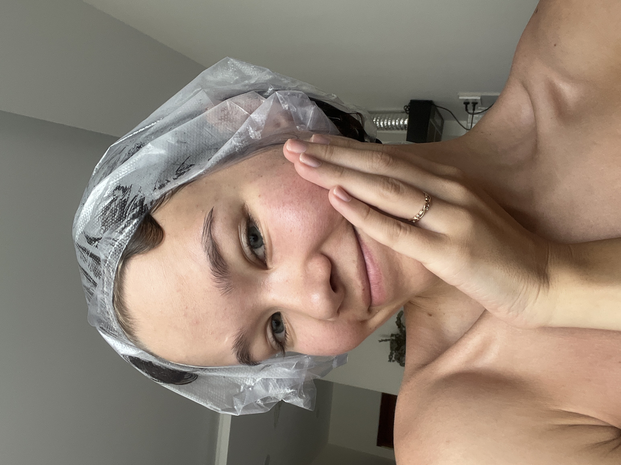 A woman wearing a clear plastic shower cap over dark hair is touching her face. She has light skin, gray eyes, and visible eyebrows. She is wearing a gold ring on her finger. The background includes a gray wall, a white ceiling, and an electrical outlet. The location is indoors. There is no indication that this image was taken in Nam Nha Trang Ward, Vietnam.