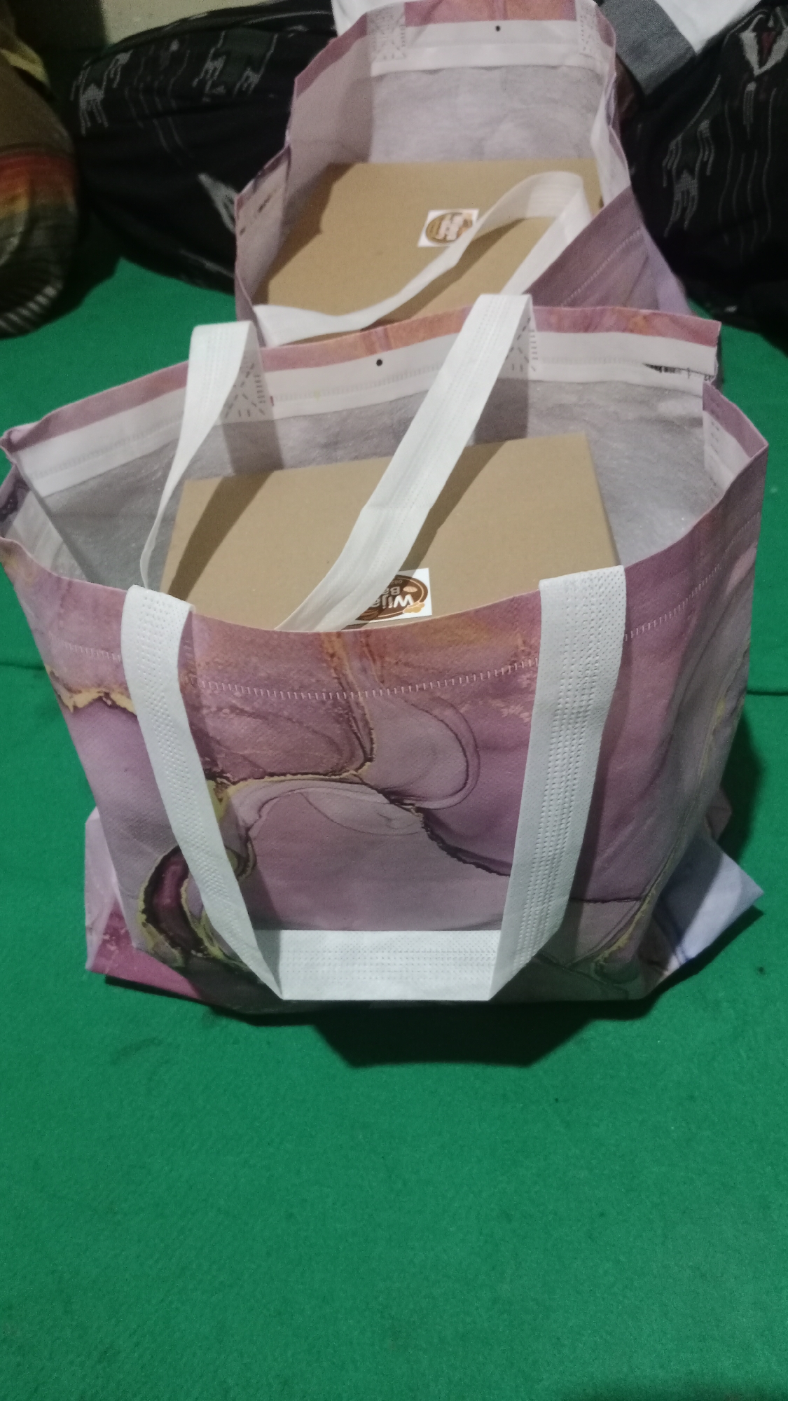 Two reusable shopping bags, one with a purple and pink abstract pattern and the other a lighter shade, are placed on a green surface, likely a tablecloth. Each bag contains a light brown cardboard box. A small, round sticker with a brown border and the letter "H" in gold is visible on the box in the top bag. A similar sticker, with the word "WIF" visible, is on the box in the lower bag. The scene is indoors.
