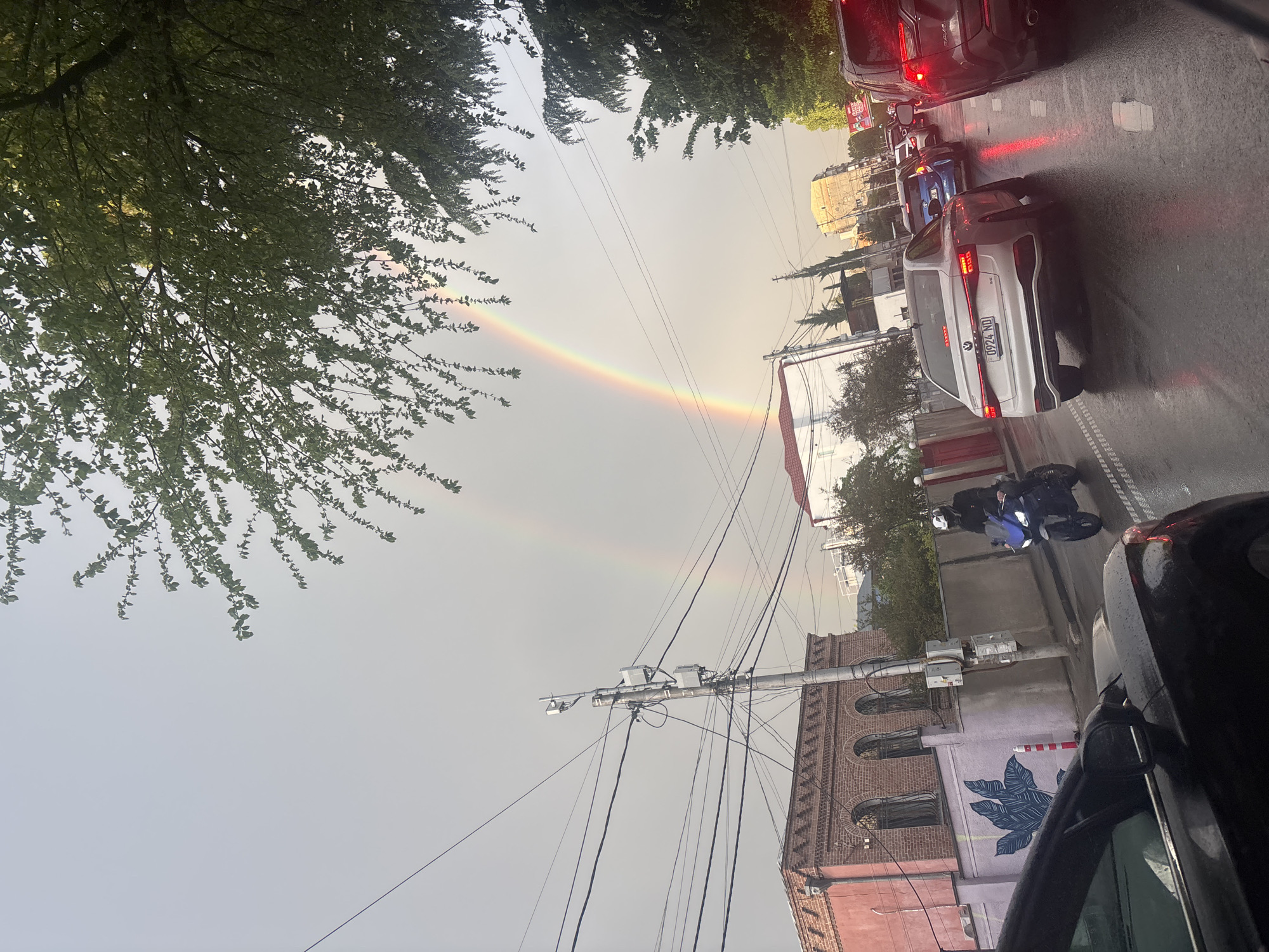 The image captures a street scene in Tbilisi, Georgia, featuring a double rainbow in the sky. The street is wet, indicating recent rainfall. Traffic is present, with several cars visible including a white car with a license plate that appears to read "0924 NO". A motorcycle is also present on the street. The architecture includes buildings with brick and pastel-colored facades, one of which features a blue leaf mural. Overhead power lines and foliage frame the scene.