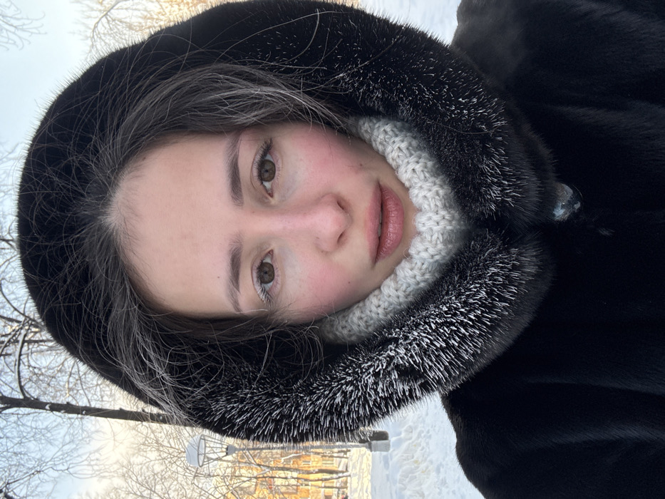 An individual, identified as female, is depicted in a close-up, chest-up portrait against a winter backdrop in Yekaterinburg, Russia. She faces forward, looking directly into the camera. Her face exhibits a flushed appearance on the cheeks and nose, consistent with cold exposure. Her eyelashes and visible hair strands under the hood show signs of white frost. She wears a dark, possibly black or dark brown, hooded coat lined with fur; the fur trim of the hood is visibly covered with white frost or ice particles. A light grey, ribbed knit neck gaiter or scarf covers her neck and lower chin. The background reveals a snow-covered environment with bare trees, also frosted, under a bright sky. A white-domed streetlamp on a dark pole and a light-colored building are visible in the distance, indicating an urban park or street setting. The scene is illuminated by daylight, suggesting a cold, clear winter day.