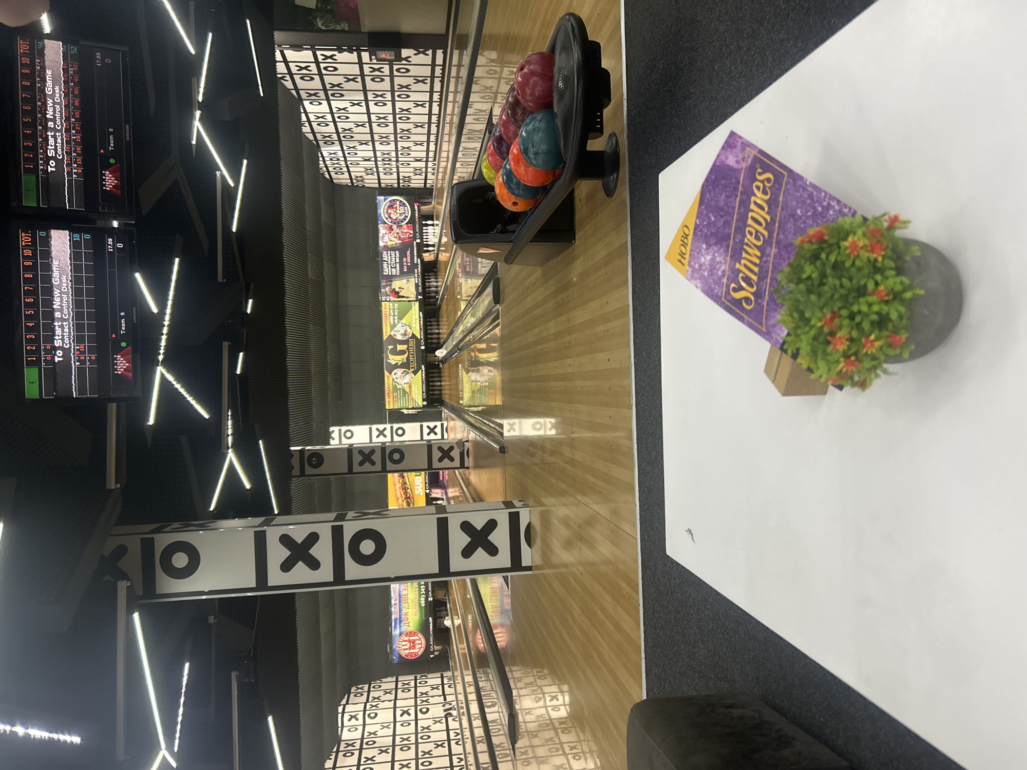 The image depicts an indoor bowling alley. Bowling lanes stretch into the distance, marked by "XOX" patterned dividers. A rack of bowling balls in various colors (red, purple, green, orange, blue) sits near the foreground, next to a table holding a Schweppes-branded item, with a small potted plant alongside it. Screens displaying scores are visible in the background. There is the logo of the Bulgarian firm "Georgievi", a sub sandwiches vendor and the sign "ДФК „ЗВЕЗД", all pointing to the location of the bowling alley as Burgas, Bulgaria. The environment appears modern with bright lights. There are no visible people. The scene appears static, showing a still moment within the bowling alley.