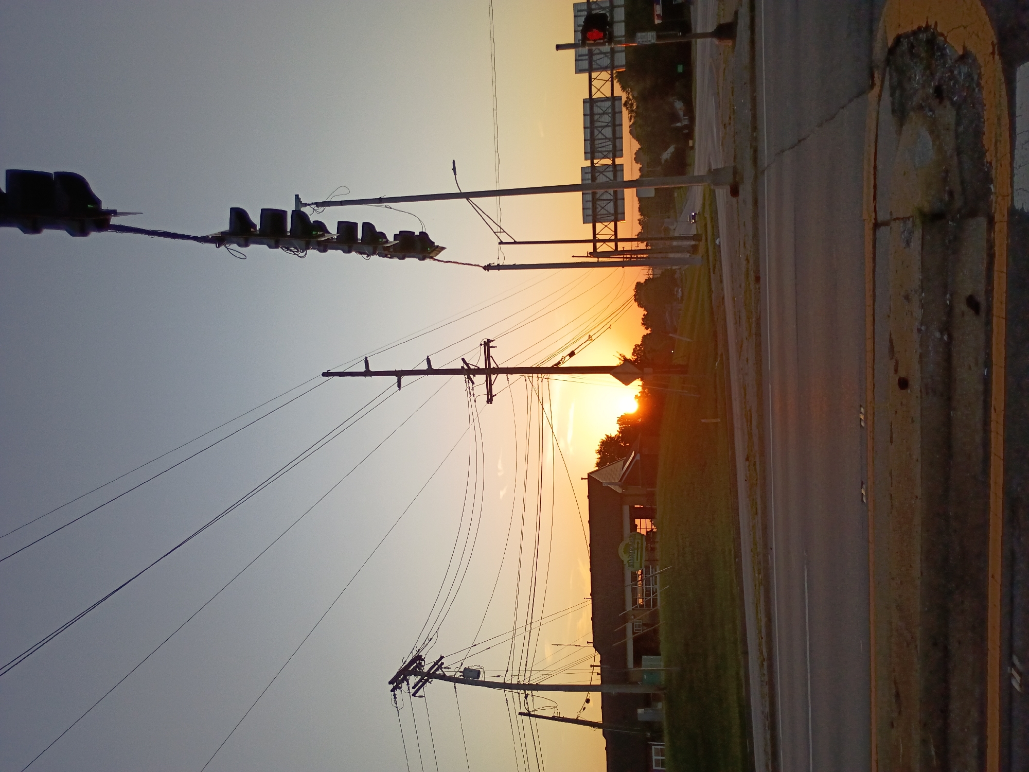 This is a vertically oriented, medium-shot, eye-level photograph of a street scene at dusk. The sky is a gradient of pale blue at the top transitioning to a bright orange-yellow near the horizon, indicating either sunrise or sunset. Silhouetted against the sky are traffic lights and utility poles with numerous wires. In the background, trees line the horizon, and a few buildings are visible, one with a green sign that appears to read "RUSTIC GRILLE." A road runs diagonally through the scene, with yellow painted lines and a cracked asphalt surface. The overall mood is peaceful and serene, with the warm light of dusk casting long shadows and bathing the scene in a soft glow. There are no people visible in the image.