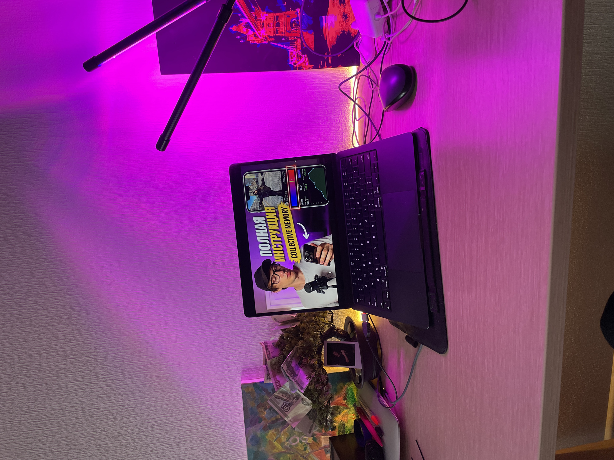 Here is a detailed description of the image:
**Location:** The image appears to have been taken indoors, possibly in a personal workspace or home office setting, in Murino, Russia.
**Objects and Subjects:**
* **Laptop:** A dark-colored laptop is positioned on a desk. The screen displays a video with a man wearing glasses and a cap. The video title is partially visible: "ПОЛНАЯ ИНСТРУКЦИЯ - COLLECTIVE MEMORY". There are charts visible within the video.
* **Desk:** The laptop rests on a desk with a light wood-grain finish.
* **Desk Lamp:** A stand with two adjustable light arms is visible in the upper left corner. It is casting a bright pink light.
* **Computer Mouse:** A black computer mouse is located to the right of the laptop.
* **Cables:** Various white cables are tangled around the desk, extending from the laptop and other devices.
* **Wall Decor:** A painting with a cityscape is hung on the wall behind the desk, as well as a money tree with Russian ruble banknotes hanging on its branches.
* **Polaroid Photo:** A Polaroid-style photo is positioned near the desk.
* **Painting:** A colorful painting is visible under the money tree.
**Environment:**
* The room is lit with a strong pink light from the desk lamp, creating an unusual color cast on the scene.
* The background features a wall with a slightly textured finish.
* The overall setting appears to be a modern and potentially creative workspace.