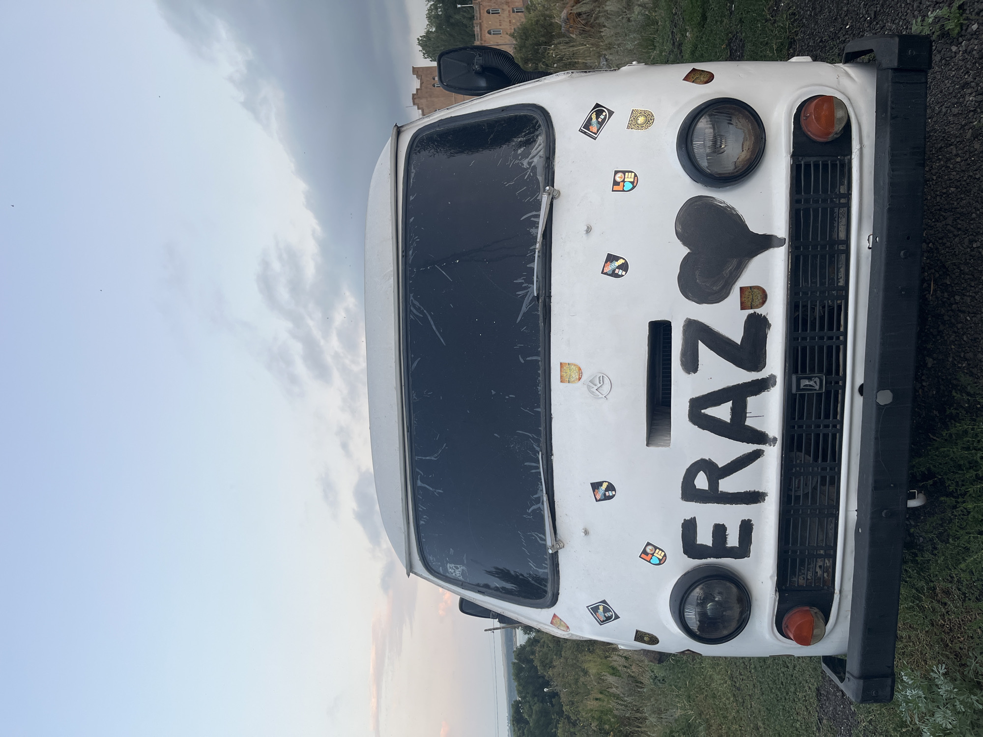 A white van is parked outdoors on a slightly elevated, grassy area. The van is the primary subject, facing the viewer. Black painted letters spelling "ERAZ" are prominent on the front of the van, accompanied by a black painted heart to the right of the lettering. Numerous stickers, many appearing to be crests or emblems, are affixed to the front of the van. The van has round headlights and a black bumper. The sky above is partly cloudy with soft, muted colors. In the background, beyond the van, a body of water and a line of trees are visible, suggesting a natural or semi-rural setting. To the right, an older, multi-story building with arched windows, possibly a historical structure, is visible amongst trees. The location is identified as Hayravanq, Armenia.