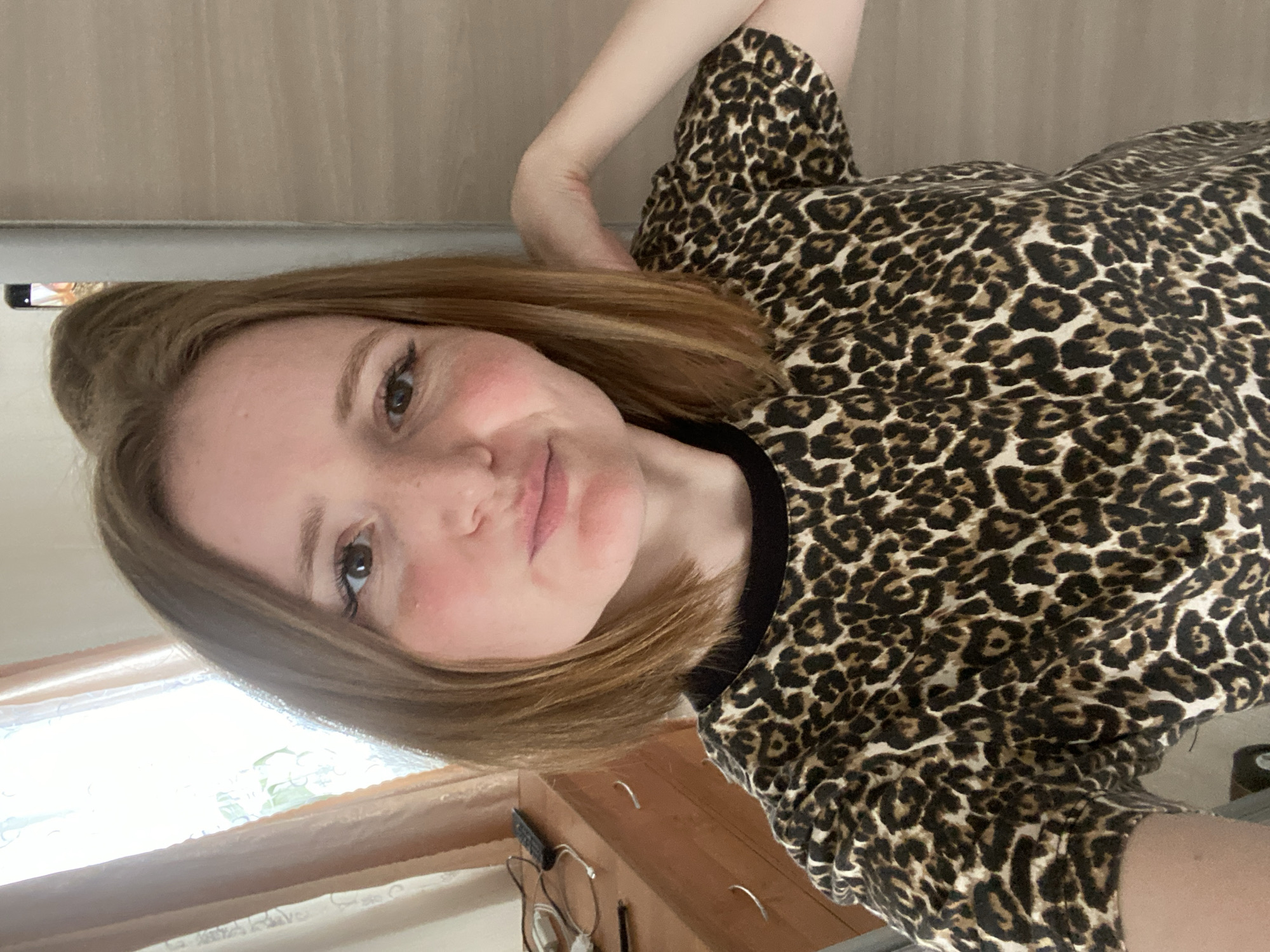 A young woman with auburn hair, wearing a leopard print t-shirt, is taking a selfie. She is indoors, with a dresser and a window with curtains visible in the background. The image was taken in Chaikovsky, Russia.