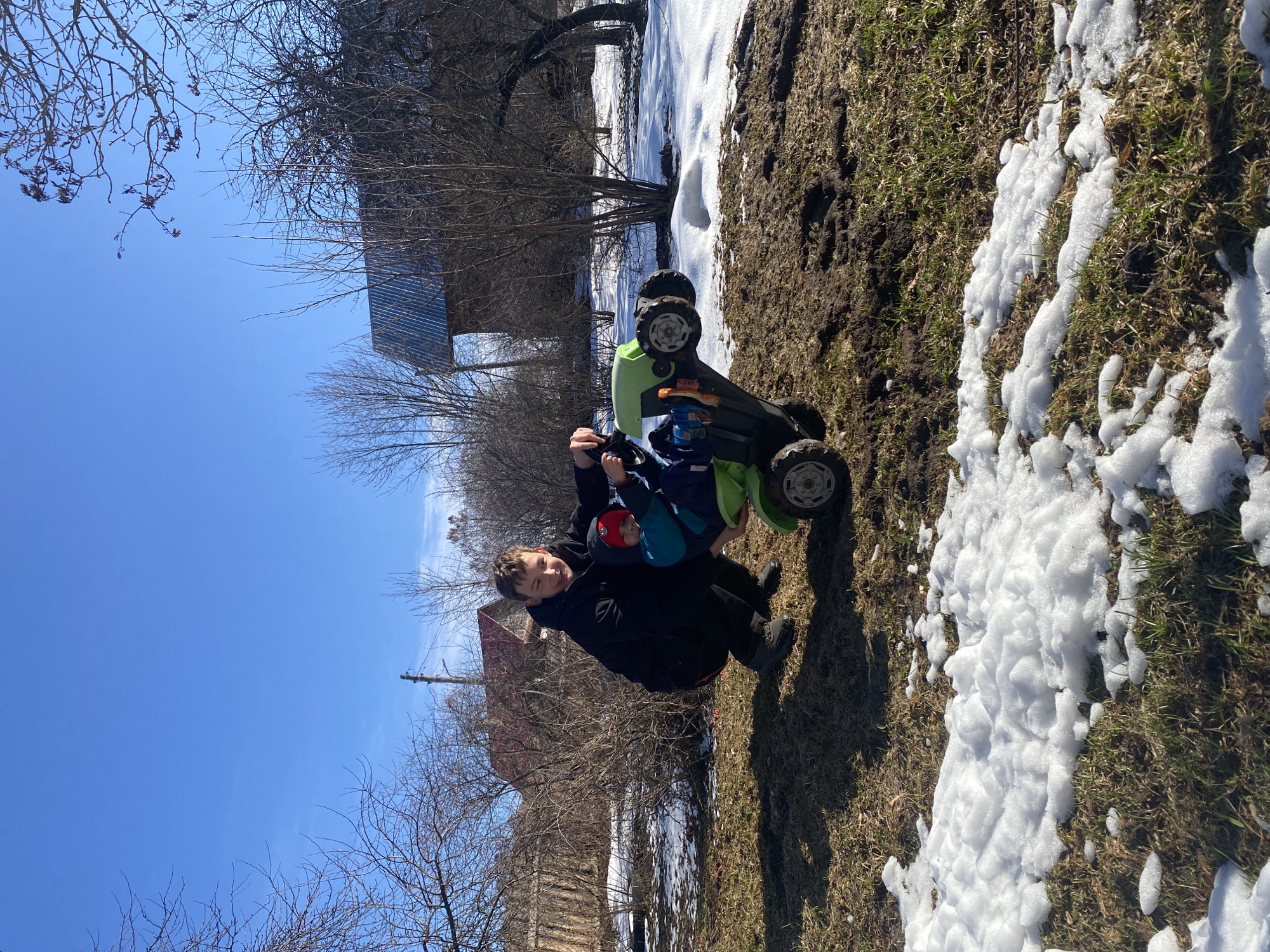The image shows two individuals in an outdoor setting. One is a boy wearing a black jacket, squatting with another child seated on a green and black toy vehicle. The ground is covered with a mix of grass, patches of snow, and bare dirt. Behind them, there are buildings, bare trees, and a clear blue sky. The location is Ольховка, Russia.