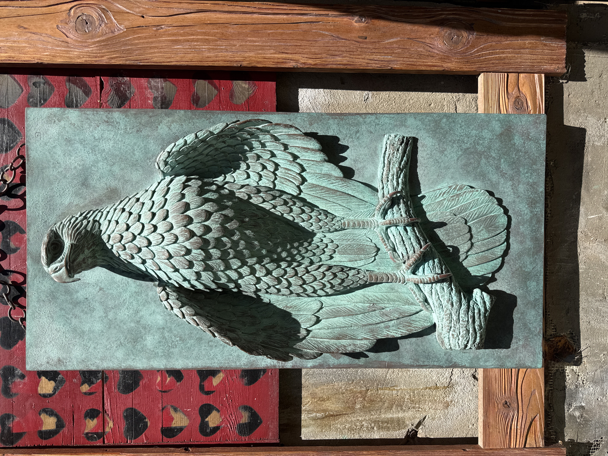 An outdoor, eye-level, medium shot captures a relief sculpture of a raptor perched on a branch, mounted on a wall. The sculpture is rendered in a verdigris patina, accentuating the detailed feathers and sharp talons. The background is a rustic wooden wall with a red panel featuring a pattern of black and gold hearts. To the right of the sculpture, a large, roughly hewn wooden beam is visible. The sculpture is likely located in Kolomyia, Ukraine, given the context. The scene is lit by natural sunlight, casting distinct shadows that highlight the three-dimensional quality of the artwork.