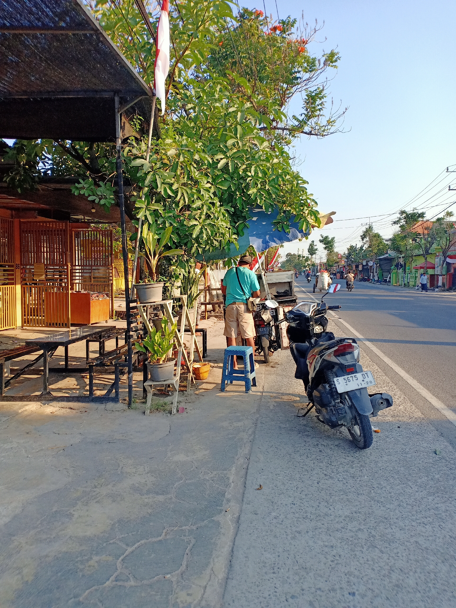 In Ngumpak Dalem, Indonesia, an outdoor scene unfolds on a sunny day. A person wearing a teal shirt and khaki shorts stands next to a food cart, which is partially obscured by a blue tarp and lush green foliage. A red and white Indonesian flag flutters nearby. In front of the food cart, a small blue plastic stool is placed on the concrete sidewalk. Two motorcycles are parked along the side of the road, one black and white with license plate S 5675 DT, and another black motorcycle. The street is lined with trees, buildings, and overhead power lines, and several other motorcycles and a few pedestrians are visible further down the road, suggesting a typical day in a populated area. The foreground features a cracked concrete walkway and a simple wooden bench.