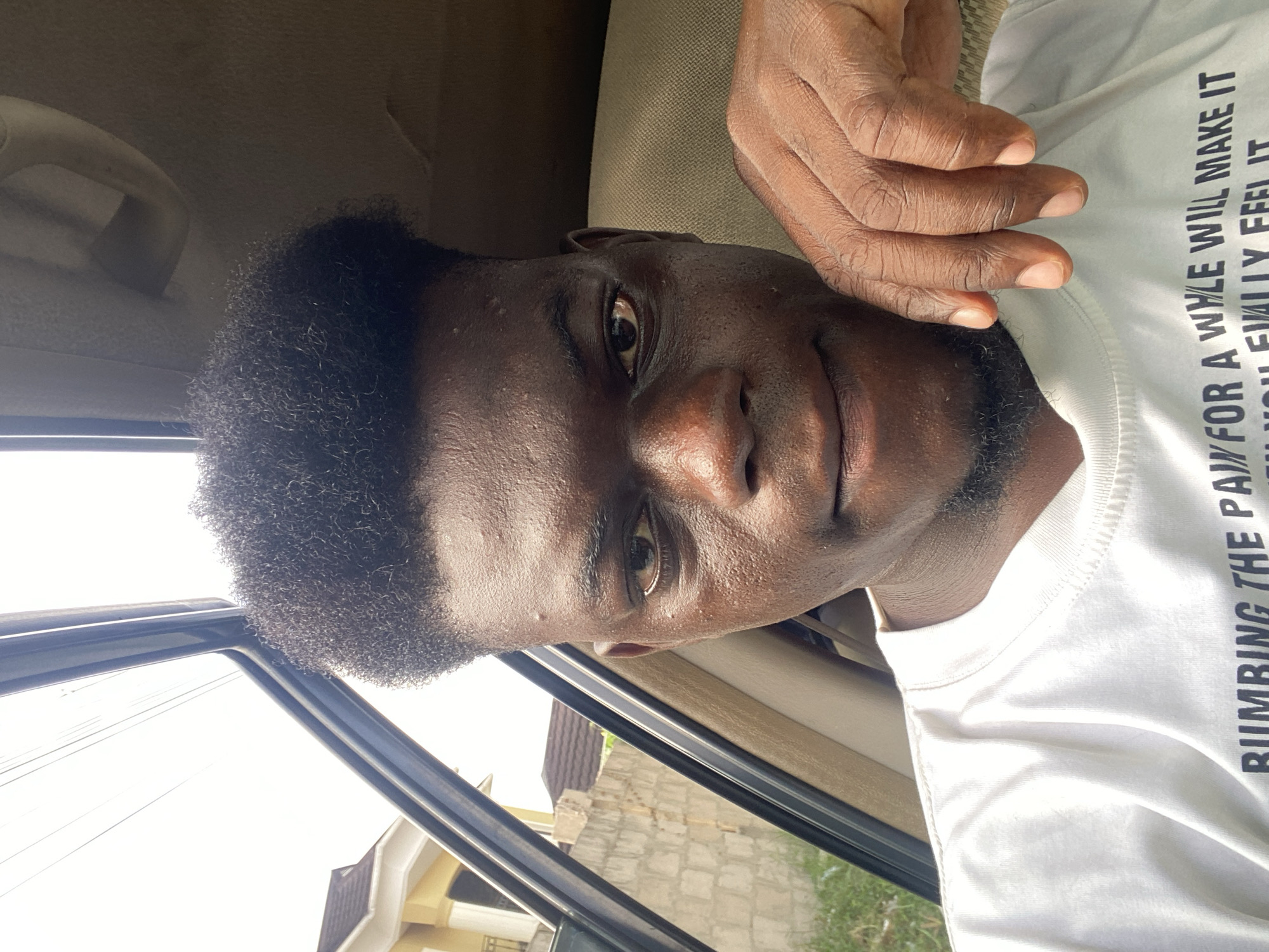 A close-up portrait shows a Black man with a dark skin tone, short curly dark hair, and a light beard or stubble. He is looking directly at the camera with a neutral expression. The image is oriented sideways, with his head positioned on the left.
He is wearing a white t-shirt, and his right hand is visible resting near his shoulder or neck area, with fingers slightly curled. The setting appears to be inside a vehicle, indicated by the car's headliner and a grab handle visible in the upper left corner. Through the car window, a bright daytime sky, utility lines, and partial views of buildings, including a house with a dark roof and a stone wall structure, are visible in the background. The bright lighting suggests it is sunny.
Visible text on his white t-shirt reads:
"RUMBLING THE PA" (with the rest of the word obscured)
"FOR A WYLE WILL MAKE IT"
"YOU EVALLY FEEL IT"