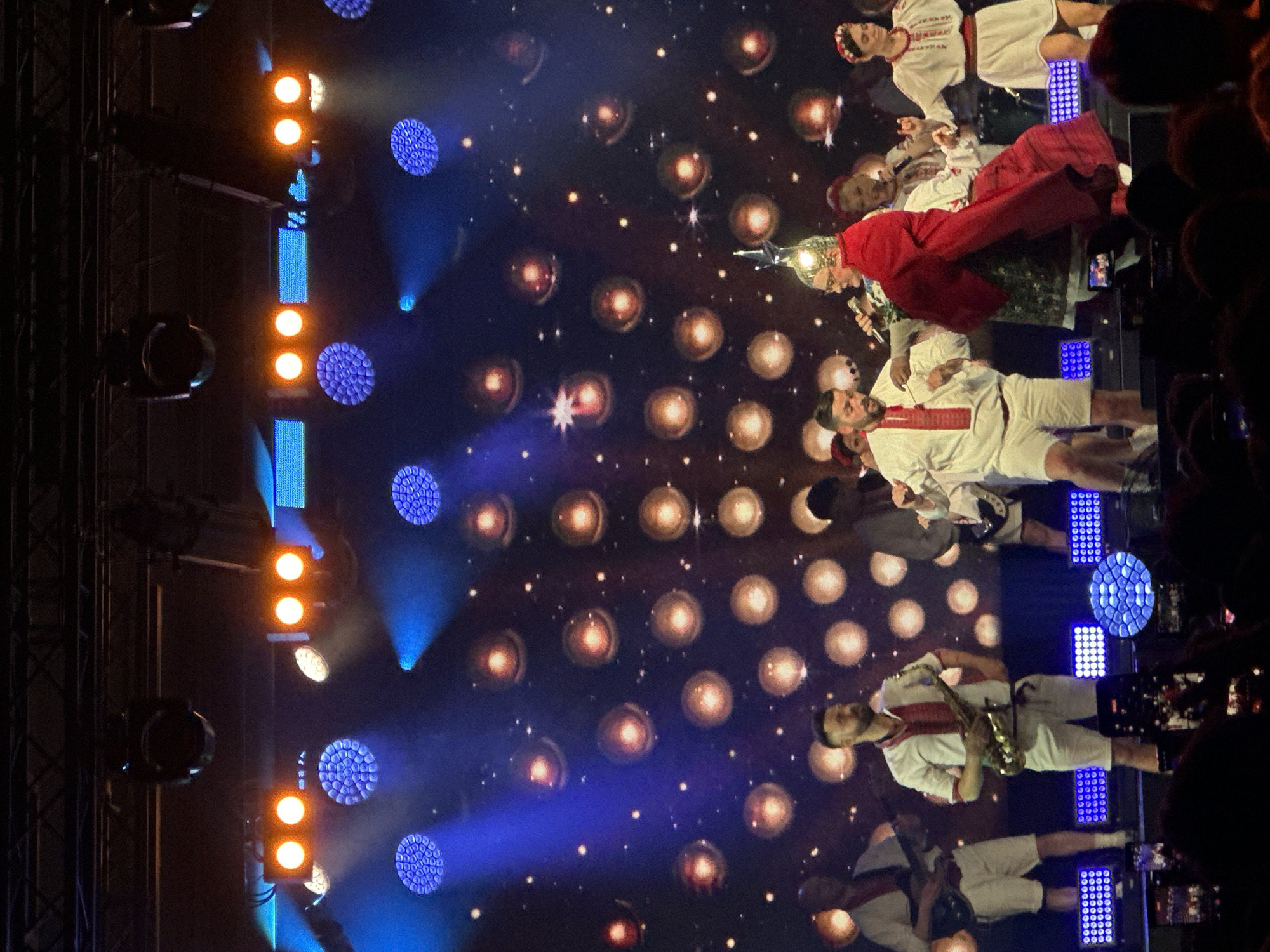 A rotated eye-level indoor shot depicts a stage with performers set against a backdrop of circular lights and stars. The event is taking place in an unknown city and region. The lighting rig on the left side of the frame features a vertical stack of orange spotlights and grid-patterned blue lights. The performers on stage are dressed in white costumes with red accents, some holding musical instruments such as a saxophone. Other subjects are singing into microphones and wearing headgear. The foreground of the shot is blurred with heads in the audience, some holding up phones, obscuring the view.