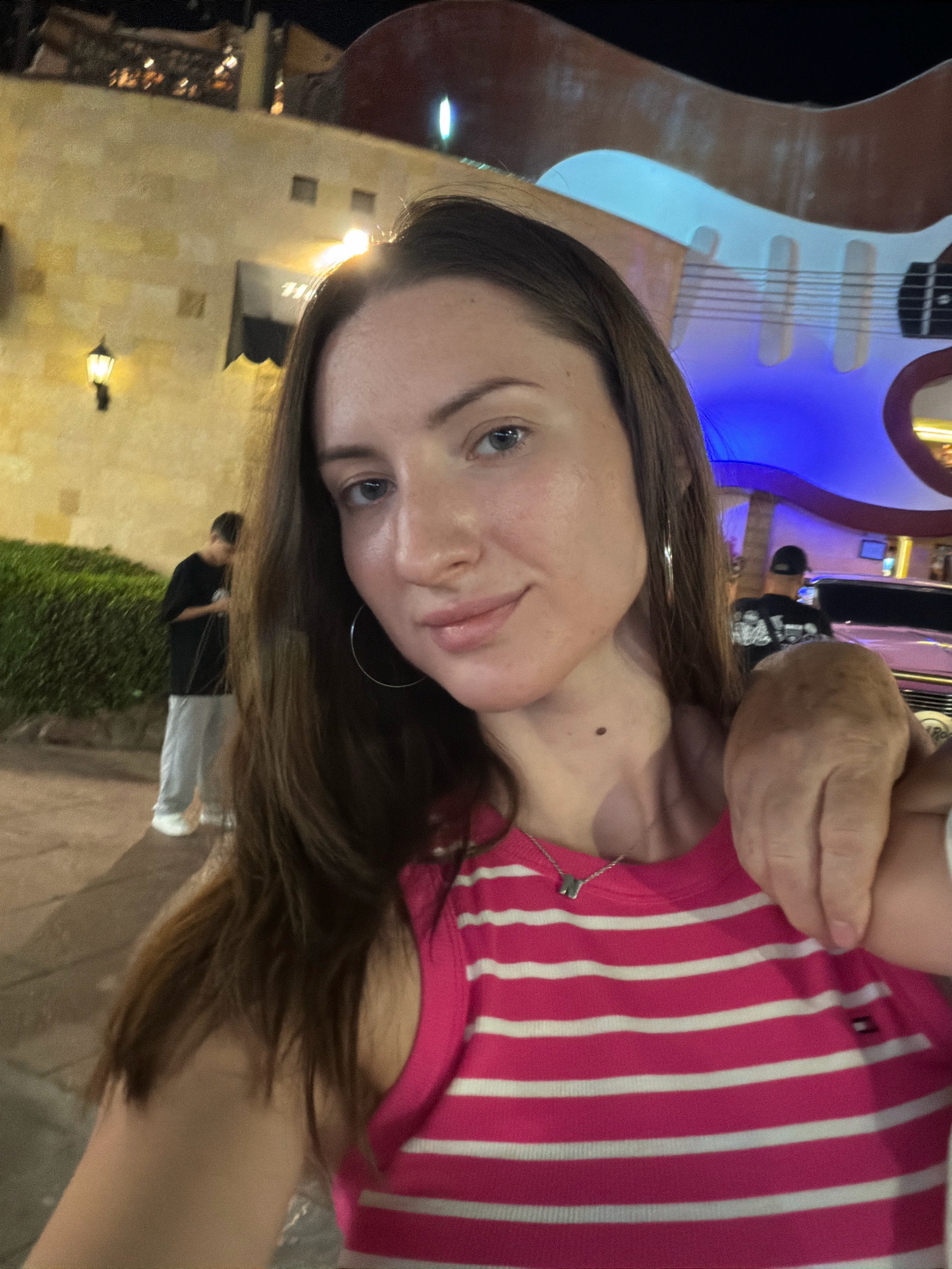 The image depicts a selfie taken at night, likely outdoors, with a prominent element of a large, illuminated guitar sculpture in the background. The location is identified as Sharm El Sheikh, Egypt, and specifically the Hard Rock Cafe, which is known for its iconic guitar sculptures and vibrant evening atmosphere.
In the foreground, a young woman with long, dark hair is looking directly at the camera. She is wearing a bright pink and white striped tank top with a small logo on the chest, and a delicate silver necklace with a charm. A person's arm is visible resting on her shoulder, partially obscuring her neck and upper chest. The woman appears to be smiling slightly.
The background reveals elements of a city environment at night. To the left, there's a building with a rustic facade, illuminated by warm-toned lights, suggesting it could be a restaurant or shop. Green bushes line a pathway in front of this building. Further in the background, two individuals are visible; one in a black t-shirt and grey pants, and another partially obscured. A pink car, possibly a limousine, is parked on the right side of the image. The overall scene is brightly lit with a mix of artificial light sources, creating a lively and somewhat festive ambiance typical of a tourist-centric location in Sharm El Sheikh. The iconic guitar sculpture, illuminated with blue and white lights, dominates the upper portion of the frame and serves as a clear indicator of the Hard Rock Cafe establishment.