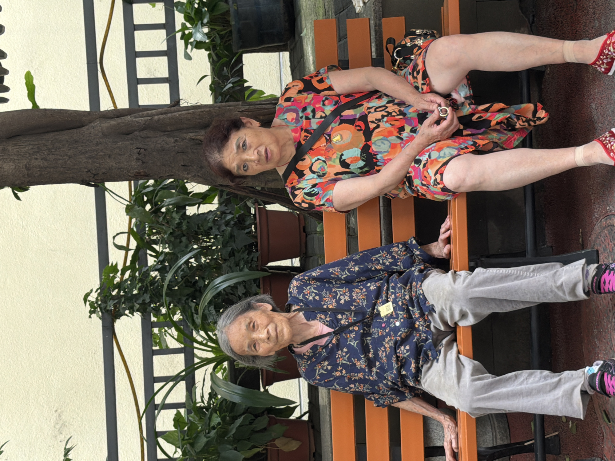 Two elderly Asian women are seated side-by-side on an orange park bench. The woman on the left has grey hair and is wearing a dark blue floral print shirt and light grey pants. She has her hands resting on her lap and appears to be looking forward. The woman on the right has dark brown hair and is wearing a colorful, abstract-patterned dress. She is holding a small gold ring in her right hand and looking slightly to her left. A black crossbody bag is slung over her shoulder, and a patterned handbag rests on the bench beside her. Behind the women, there are potted plants and a large tree trunk. The background wall is a light yellow color, and a black metal fence is visible. The setting appears to be outdoors, in a public area within Yuzhong District, China.