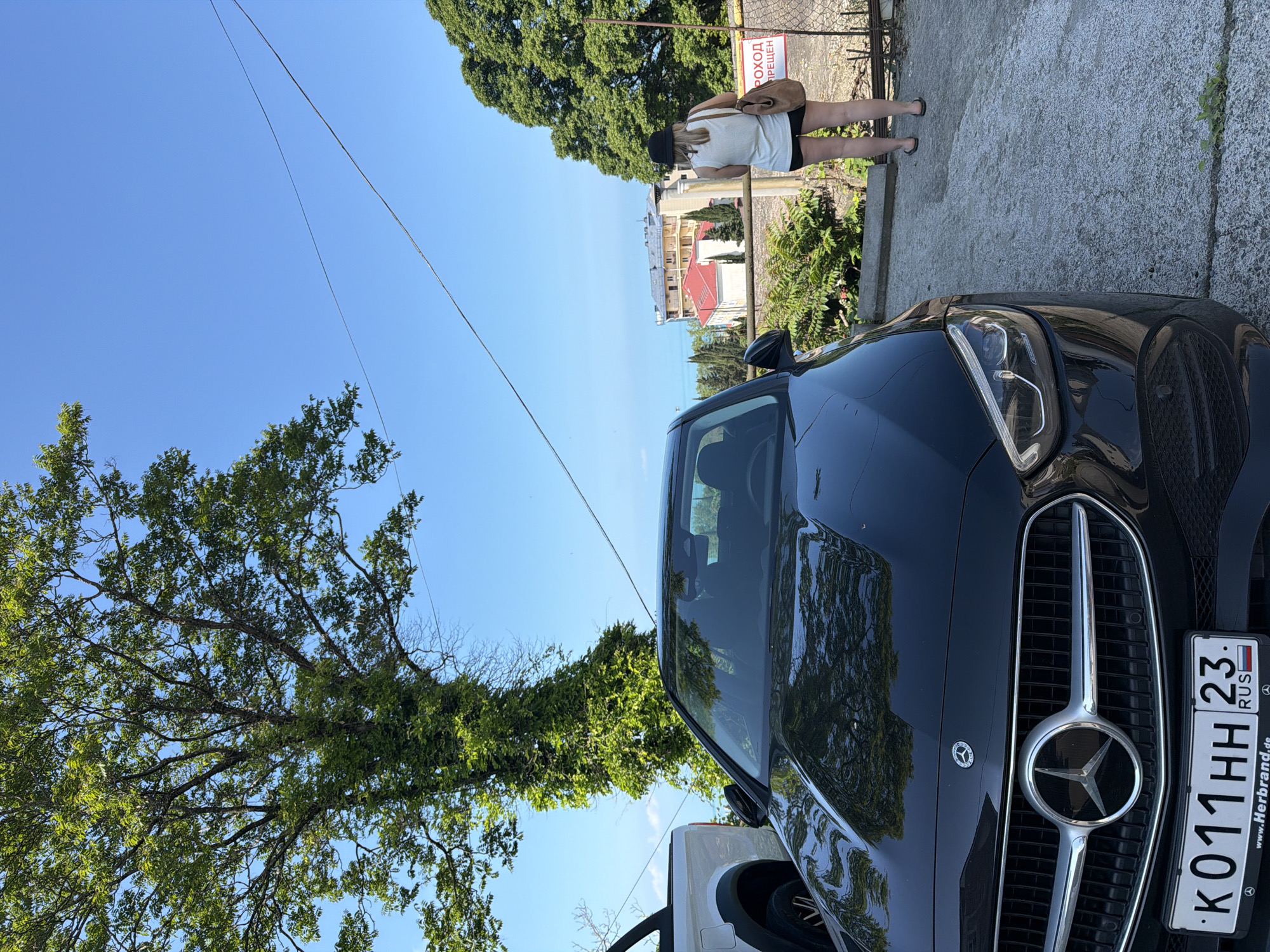 In Sochi, Russia, a black Mercedes-Benz sedan is parked on a concrete surface near a fence with a sign that reads "ВХОД ВОСПРЕЩЕН" (Entrance Forbidden). A woman wearing a white top, black shorts, and a hat is standing near the fence, facing away from the camera, and carrying a brown bag over her shoulder. The car's license plate is visible and reads "K011HH 23". To the left of the Mercedes, the rear of a white pickup truck is partially visible. Lush green trees, including a large tree with abundant foliage covering its trunk, are prominent in the background, with the blue sky and a glimpse of the sea or ocean visible in the distance beyond some buildings.