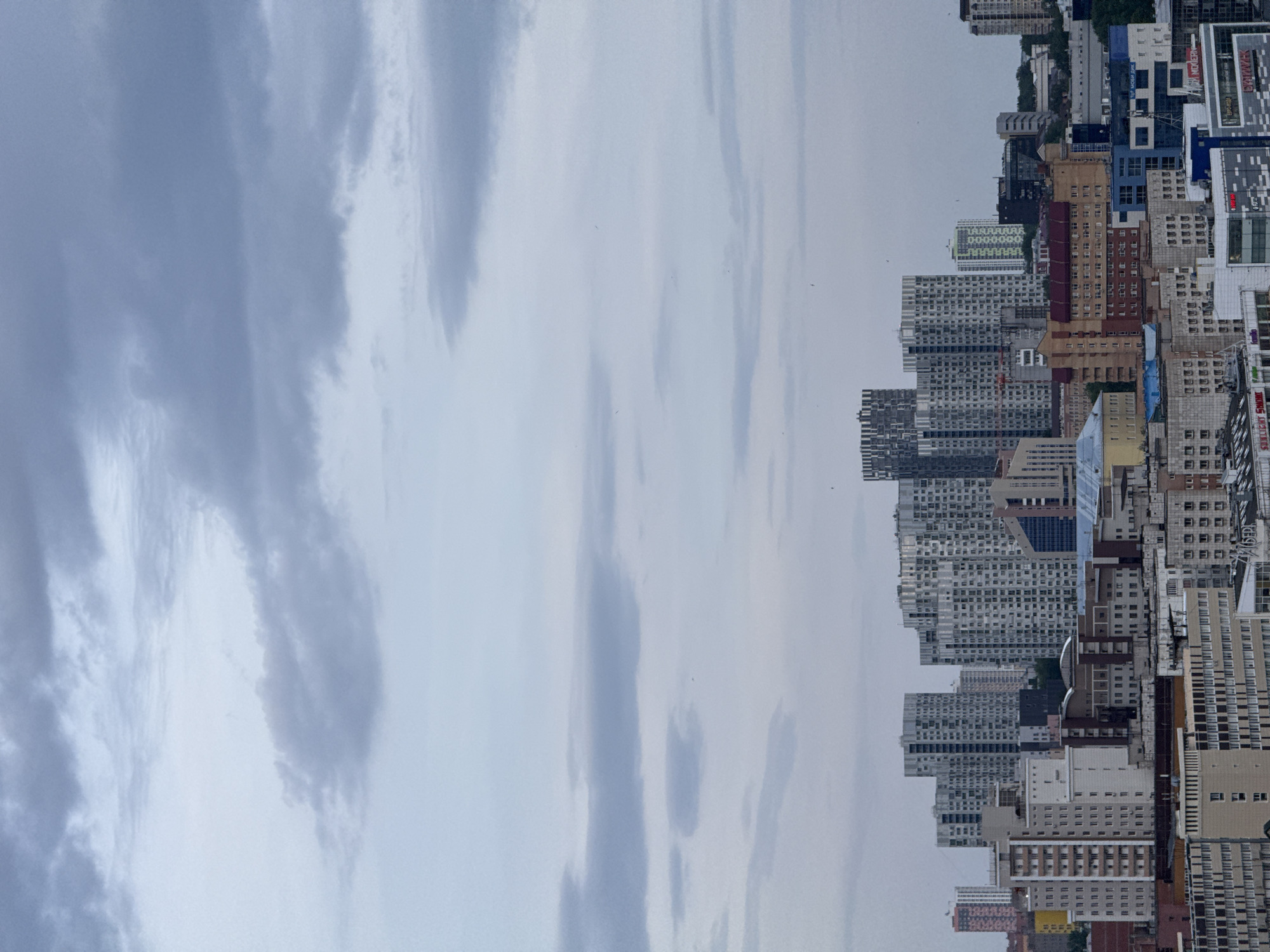The image shows a cityscape of Perm, Russia, viewed from an elevated position. Tall apartment buildings and commercial structures dominate the foreground, rising against a backdrop of a cloudy sky. The buildings vary in color and design, with a mix of gray, brown, and blue facades. The sky features wispy cloud formations with hints of blue peering through. The picture is rotated 90 degrees counter-clockwise.