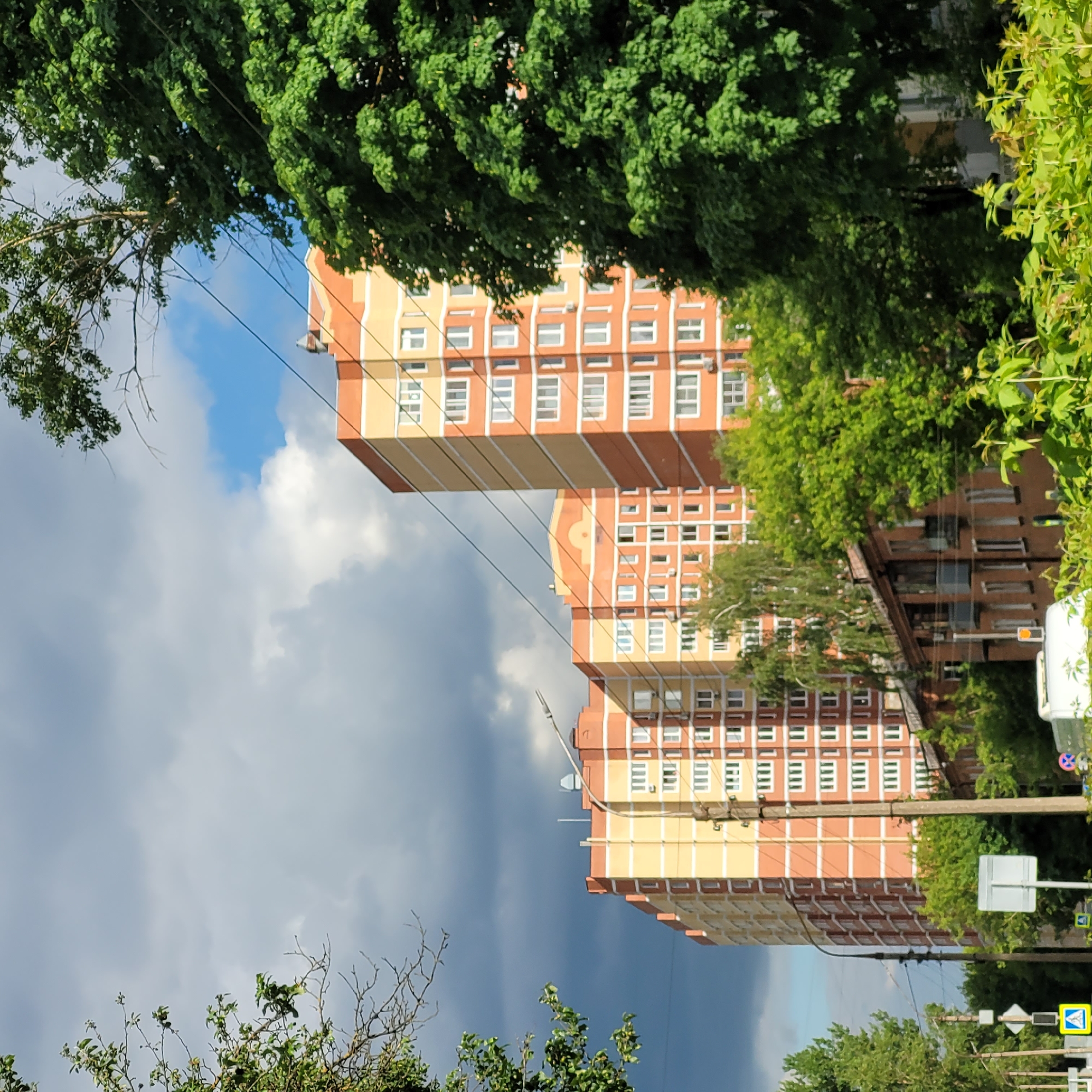 The provided image, which appears to be a landscape photograph rotated 90 degrees counter-clockwise, captures an urban or suburban street scene on a day with mixed weather.
In the center of the frame, if viewed upright, stand two tall, multi-story residential buildings, likely apartment blocks. They feature a distinct facade of alternating orange-red and light yellow or beige sections, with numerous white-framed windows. The building on the left is partially obscured by trees.
Lush green trees and foliage frame the top and bottom (left and right, respectively, in the rotated image) of the scene, partially obscuring the buildings and the sky. Bare tree branches are also visible among the green leaves. Several utility lines or cables stretch across the sky and in front of the buildings.
The sky is a notable element, displaying both bright blue patches with white clouds and a significant portion of dark, heavy grey clouds, suggesting dynamic or approaching stormy weather.
On the far right side of the image (top right if rotated upright), behind some foliage, several elements of street infrastructure are visible: a white utility box or post, a yellow traffic light (appearing amber), and multiple road signs. One clearly visible sign is a yellow diamond-shaped pedestrian crossing sign with a black symbol. Other signs, including a white square sign and a circular red-bordered sign, are partially obscured.
No specific activity is taking place; the scene is static. The time of day is daytime, evidenced by the bright light on the foliage and buildings, despite the presence of dark clouds. No discernible text is visible on any objects or signs.