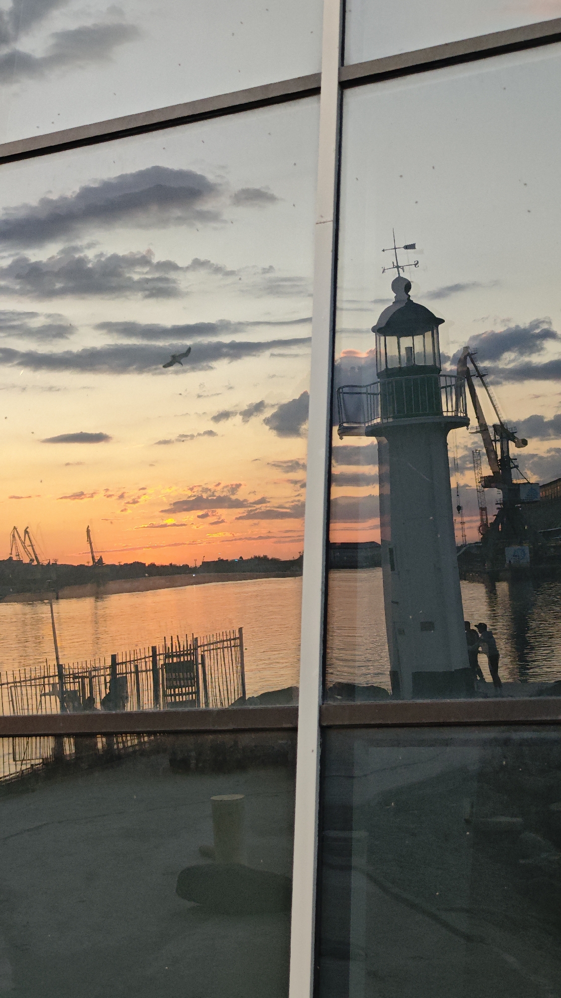 This photograph shows a view through a window reflecting a coastal scene. The window's frame divides the view. On the left is a sunset over water, with a silhouetted bird in flight and a fence along the shore. On the right, reflected in the window, is a lighthouse with a weather vane, cranes, and two figures standing on what appears to be a dock or pier. The scene likely depicts the lighthouse at the Port of Burgas, Bulgaria.