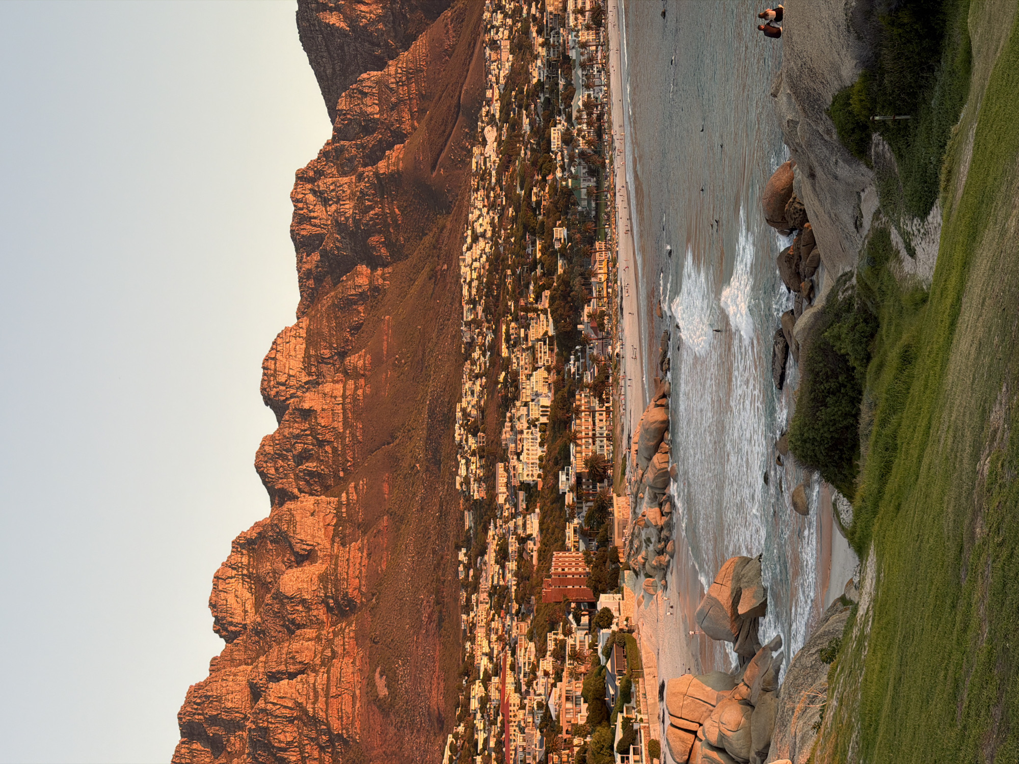 The image displays a panoramic coastal landscape at golden hour, likely sunset, in Cape Town, South Africa.
Oriented correctly, a vast, rugged mountain range dominates the left side of the frame, bathed in warm, reddish-orange light that accentuates its rocky textures and deep crevices. At the foot of these mountains and stretching towards the ocean lies a densely populated coastal town. Numerous light-colored buildings, interspersed with green trees and vegetation, create a vibrant urban tapestry glowing under the warm light.
A sandy beach forms the transition between the town and the sea. Gentle waves with white foam can be seen breaking near the shore. Small figures are visible on the beach, walking, and in the water, potentially swimming or surfing.
In the foreground, to the right, a grassy slope descends towards a rocky shoreline. Large, smooth boulders, ranging in color from light tan to grey, are scattered along the water's edge and on the sand. Two indistinct figures are discernible on the grassy slope.
The sky is clear, displaying a soft gradient from a pale blue at the top to a warm, light yellow closer to the horizon, reflecting the late afternoon or early morning sun. The overall scene conveys a sense of picturesque tranquility and natural grandeur. No visible text is present.