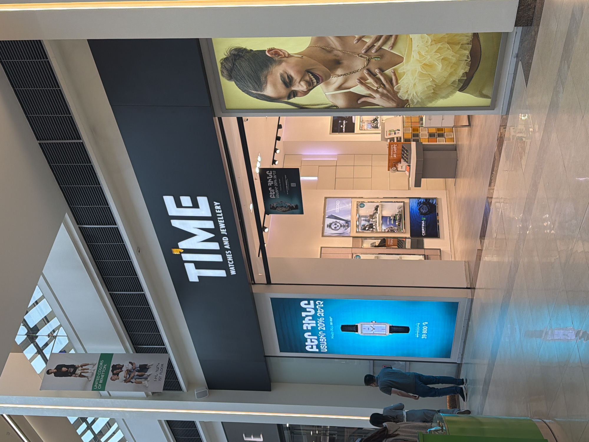 The image shows the facade of a "TIME Watches and Jewellery" store in a shopping mall. The store is located in Yerevan, Armenia.
The store front includes large advertisements above the store entrance. The advertisement on the right features a laughing woman wearing jewelry. The advertisement below it contains text in Armenian.
The store's entrance is visible, with a display of watches and jewelry inside. A man in a grey shirt walks past the storefront. Another person is sitting on the ground near a green object. Other shoppers are visible in the distance.
There is an advertisement for "UNITED COLORS OF BENETTON" on the left.