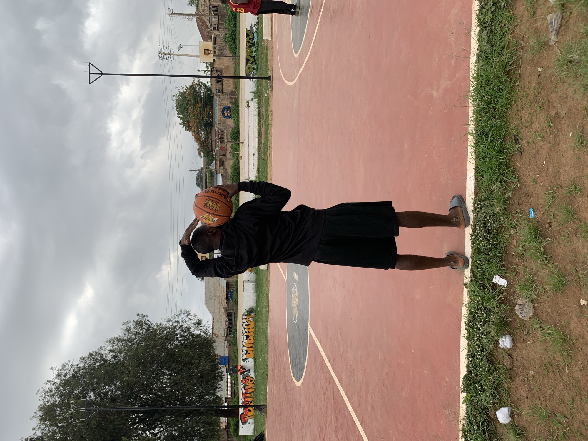 A person stands on a red-surfaced basketball court in Gura Top, Nigeria, preparing to shoot a basketball. The ball is brown with "NBA" and "LAKERS" markings. The person wears a black shirt and black shorts, with grey flip-flops. A basketball hoop and utility pole are visible in the background. A second person is seen further down the court. The court's sideline is bordered by grass and dirt with scattered debris. Graffiti is visible on a retaining wall adjacent to the court. The sky is overcast with grey clouds.