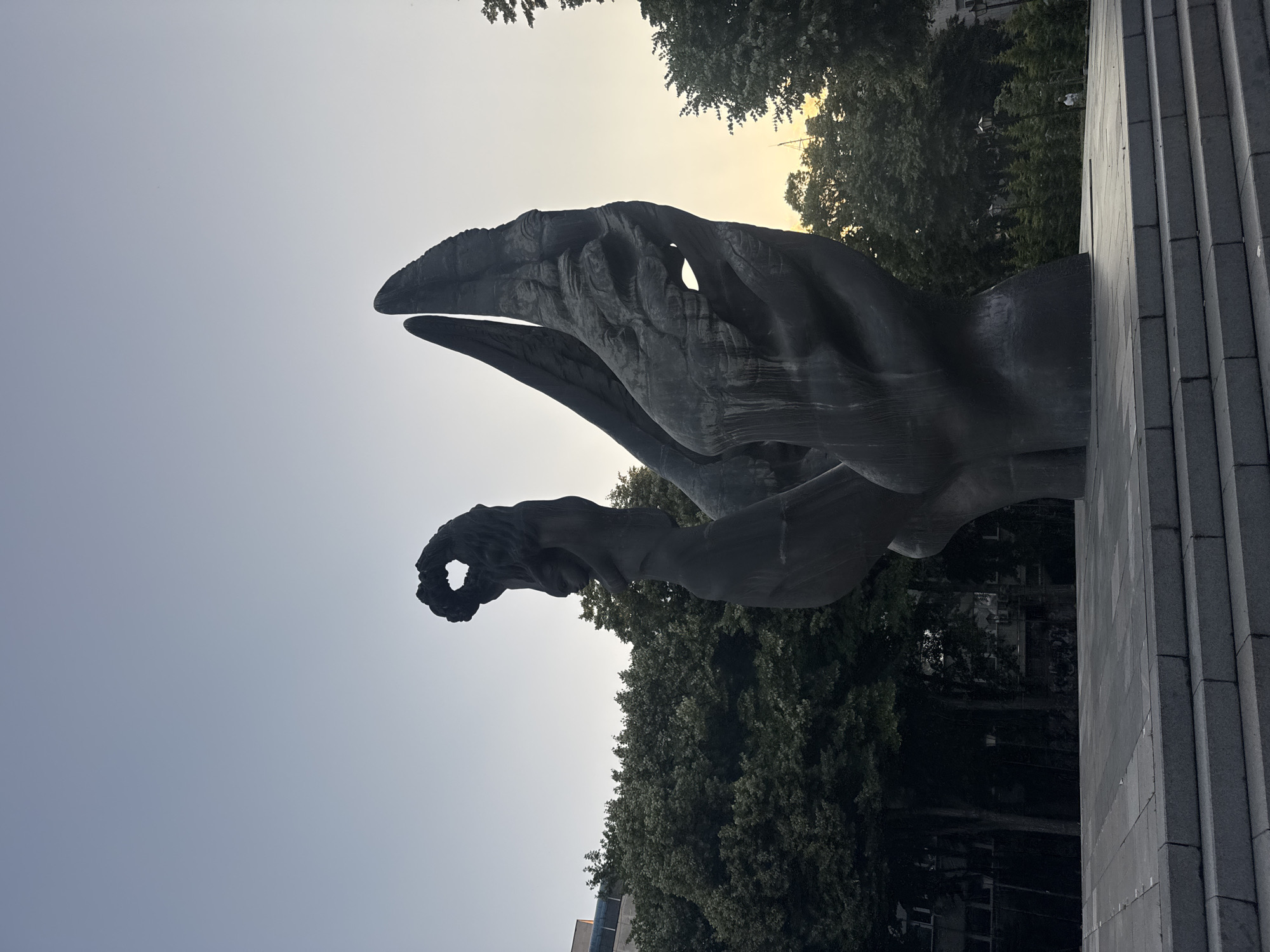 The image displays a large, dark-toned monument, identified as the Monument to the Liberators (also known as Mother Bulgaria), situated in a square near the Central Post Office in Plovdiv, Bulgaria. The monument consists of two primary sculptural elements crafted from what appears to be dark bronze or stone. On the left, a stylized female figure stands vertically, holding a wreath-like object above its head, which forms a heart-shaped aperture. To its right, a larger, abstract form rises, characterized by a textured, organic surface with wing-like or flame-like elements and irregular voids.
The monument rests on a multi-tiered base of light gray stone steps, leading up to a paved platform of similar material. The sky above transitions from a pale blue on the left to a faint yellowish-orange tint towards the right, suggesting either dawn or dusk. A dense collection of dark green trees provides a natural backdrop behind and around the monument. Portions of urban buildings are visible through the trees on the left side of the frame. In the far right background, a single individual, dressed in light attire, is seated on a low structure amidst sparse foliage. The scene is illuminated by natural light, casting the monument in a somewhat shadowed appearance, contrasting with the brighter sky.