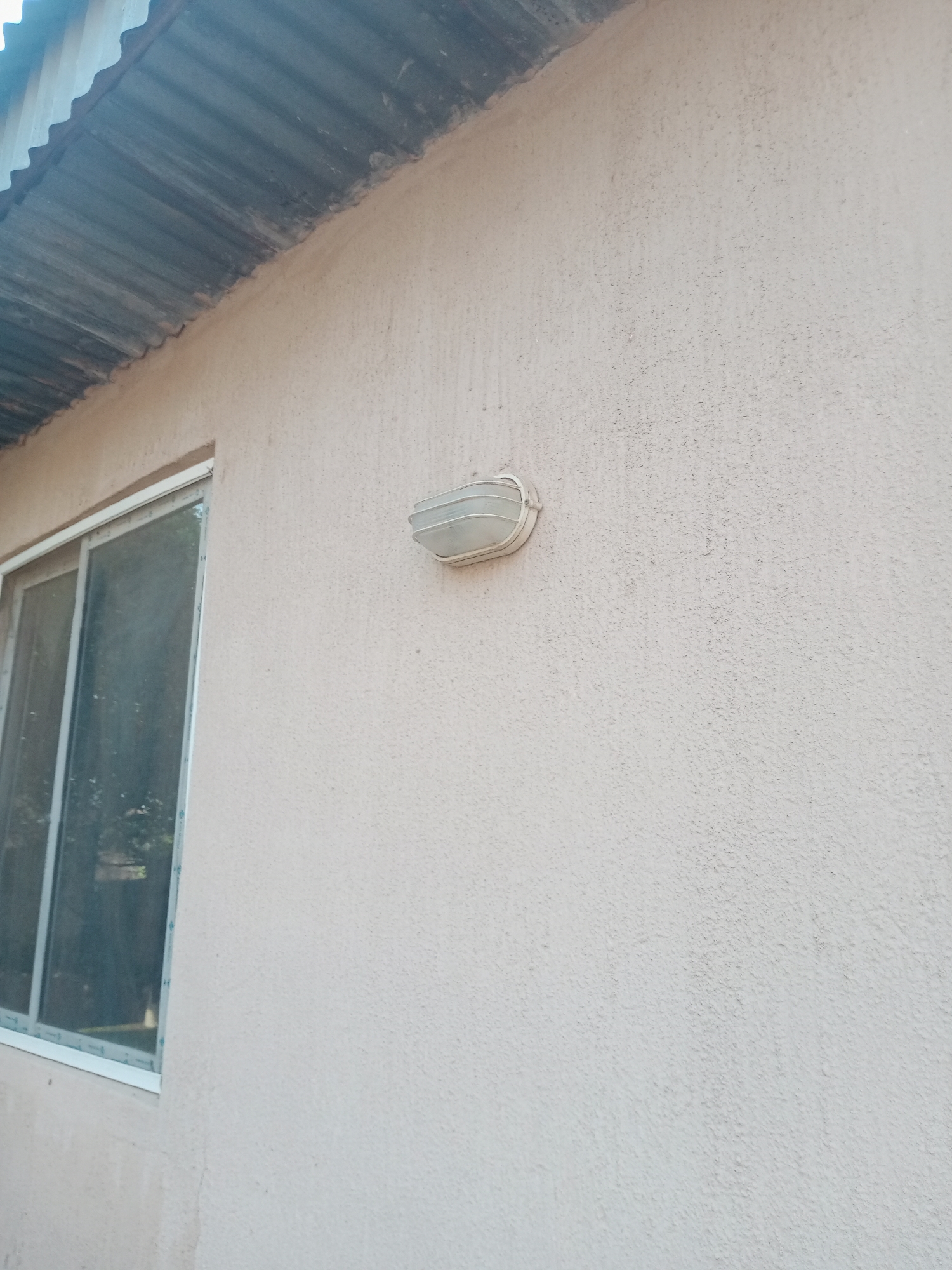 The image shows the exterior wall of a building with a corrugated metal roof. A window with a white frame and reflective glass is visible on the left side of the image. A white outdoor light fixture is mounted on the wall to the right of the window. The wall has a textured, light-colored finish. The scene appears to be outdoors in daylight, with no people or other distinguishing environmental features visible in this frame. The building's architecture and materials suggest it could be located in a residential or commercial area in Jalingo, Nigeria.