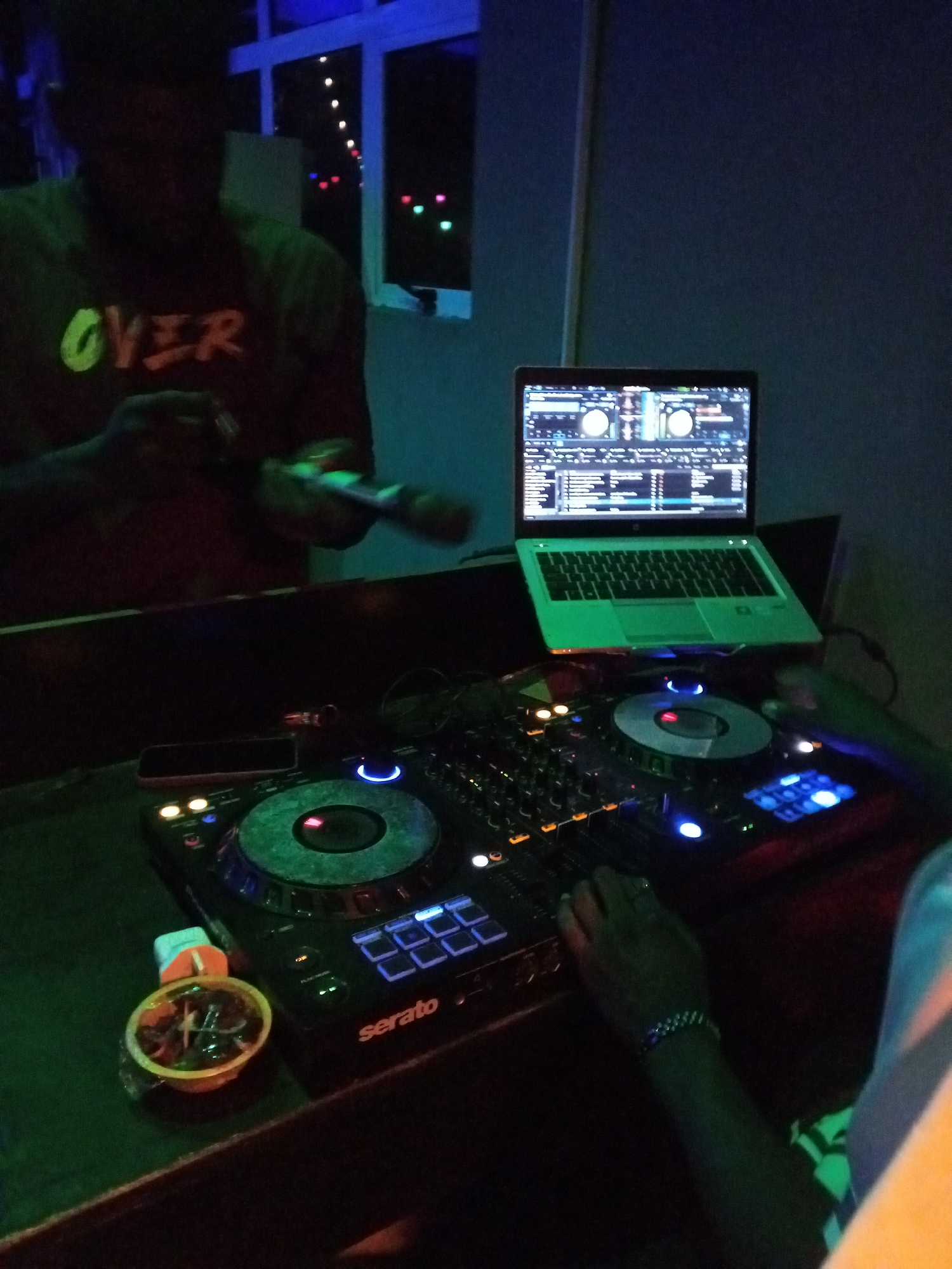 In an indoor setting with dim lighting, a person is seen from the chest up, wearing a dark shirt with the word "OVER" in a neon green font. They are holding a microphone in their right hand. In front of them, a DJ setup is visible, including a laptop displaying music software, a DJ controller with glowing blue lights and buttons, and a circular turntable. A small container with food or snacks is on the left side of the DJ controller. The background shows a window with colorful lights outside and a dark blue wall. The overall scene suggests a music performance or DJ session.