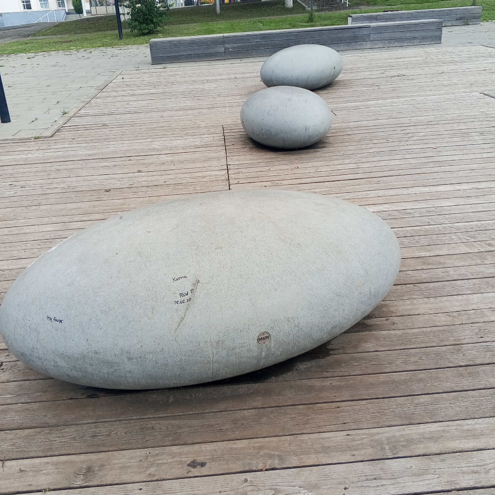 In a city park in Dyurtyuli, Russia, three large, smooth, light-grey, oval-shaped concrete benches are arranged on a wooden deck. The largest bench is in the foreground, with two smaller benches positioned behind it. The benches are weathered and have some graffiti inscribed on them. The largest bench has the inscriptions "Stray Kids", "Karma", "RU II" and "20.06.20" visible. A small circular metallic emblem with the word "DESSO" is also attached to the largest bench. The wooden deck is laid out in parallel planks. In the background, there is a paved area with some greenery and buildings.