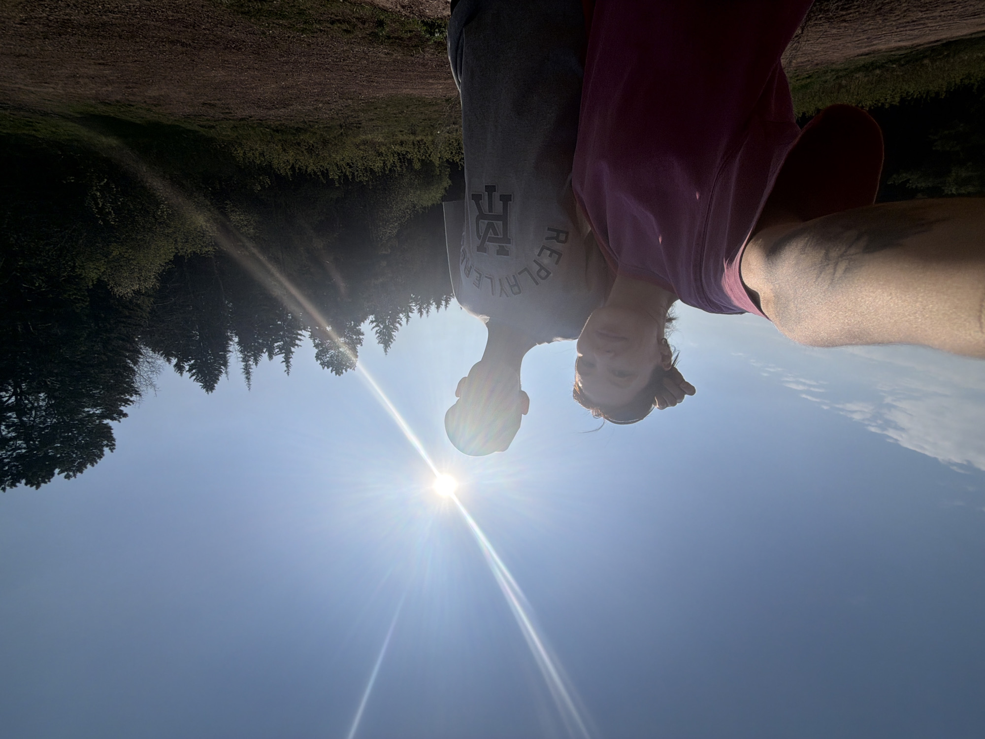 Two people are standing in an outdoor environment, possibly a park or forest area on a sunny day. The sky is clear blue, with bright sunlight. Trees are visible in the background, suggesting a natural setting. The person on the left is wearing a gray shirt with a "REPLAYLER" design. The other person is wearing a pink shirt. The location cannot be determined as Perm, Russia, with the provided information.