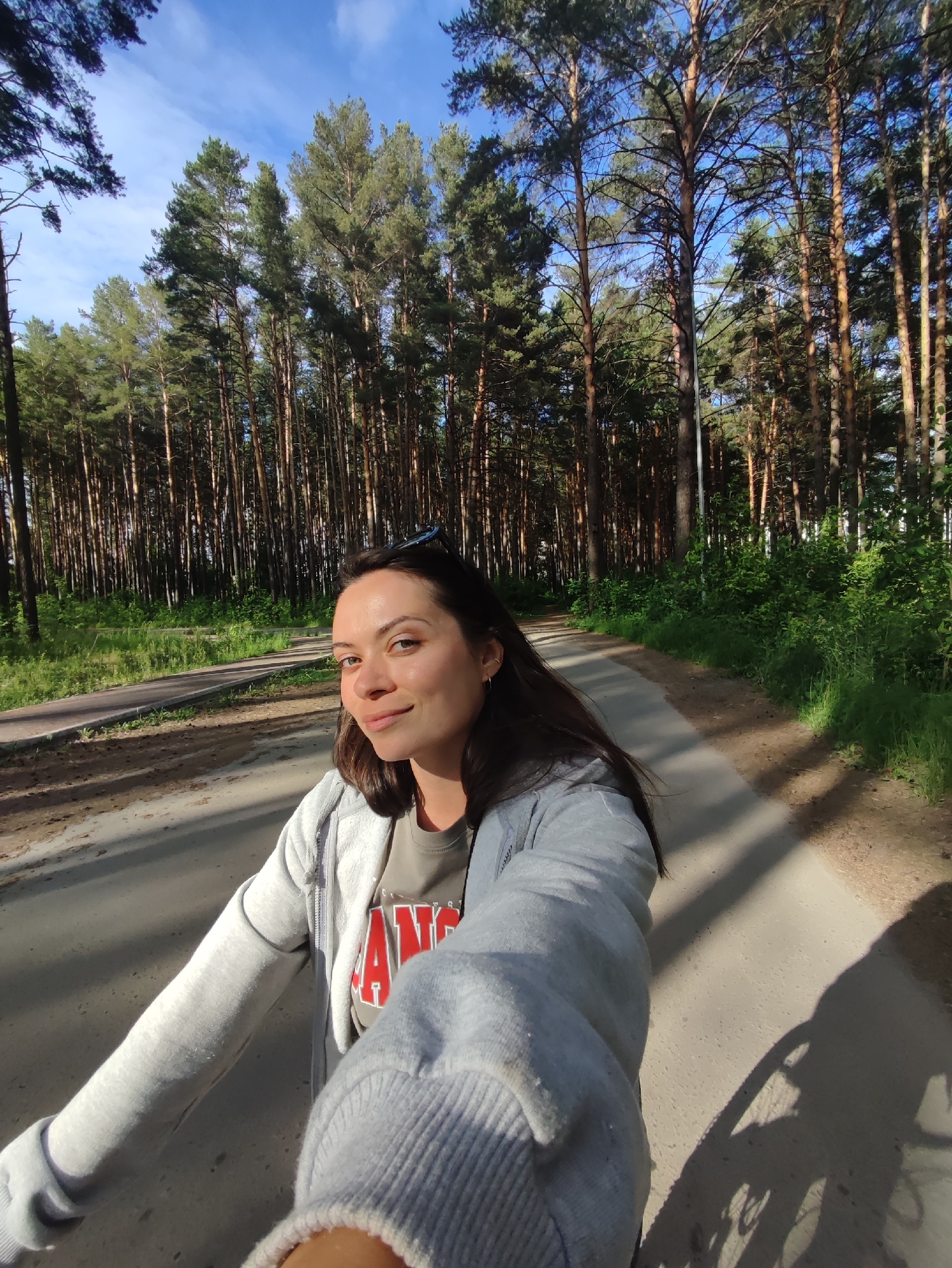 The image shows a young woman with dark hair taking a selfie while on a paved path in a pine forest. She is wearing a grey zip-up hoodie over a t-shirt with red lettering. Her left arm is extended to capture the photo. The path curves to the left in the background, and is bordered by dense pine trees on the right and more sparse trees and green foliage on the left. The sky is blue with scattered clouds. The lighting suggests it is late afternoon, casting long shadows across the path. The location is the pine forest park in the city of Chaikovsky, Russia, a popular area for recreation.